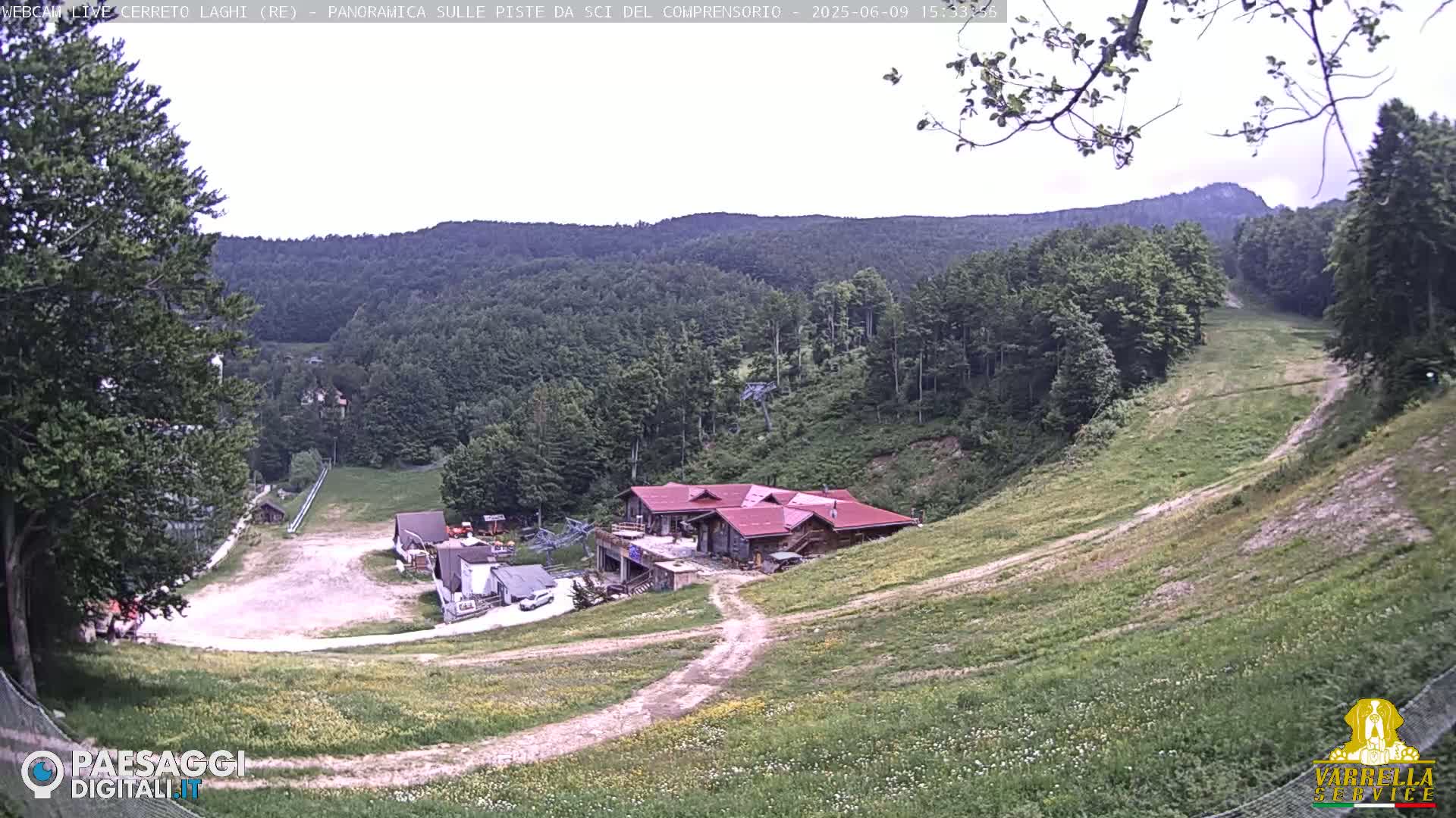 A mostly sunny view of a grassy ski slope with a few buildings and forested hills in the background.