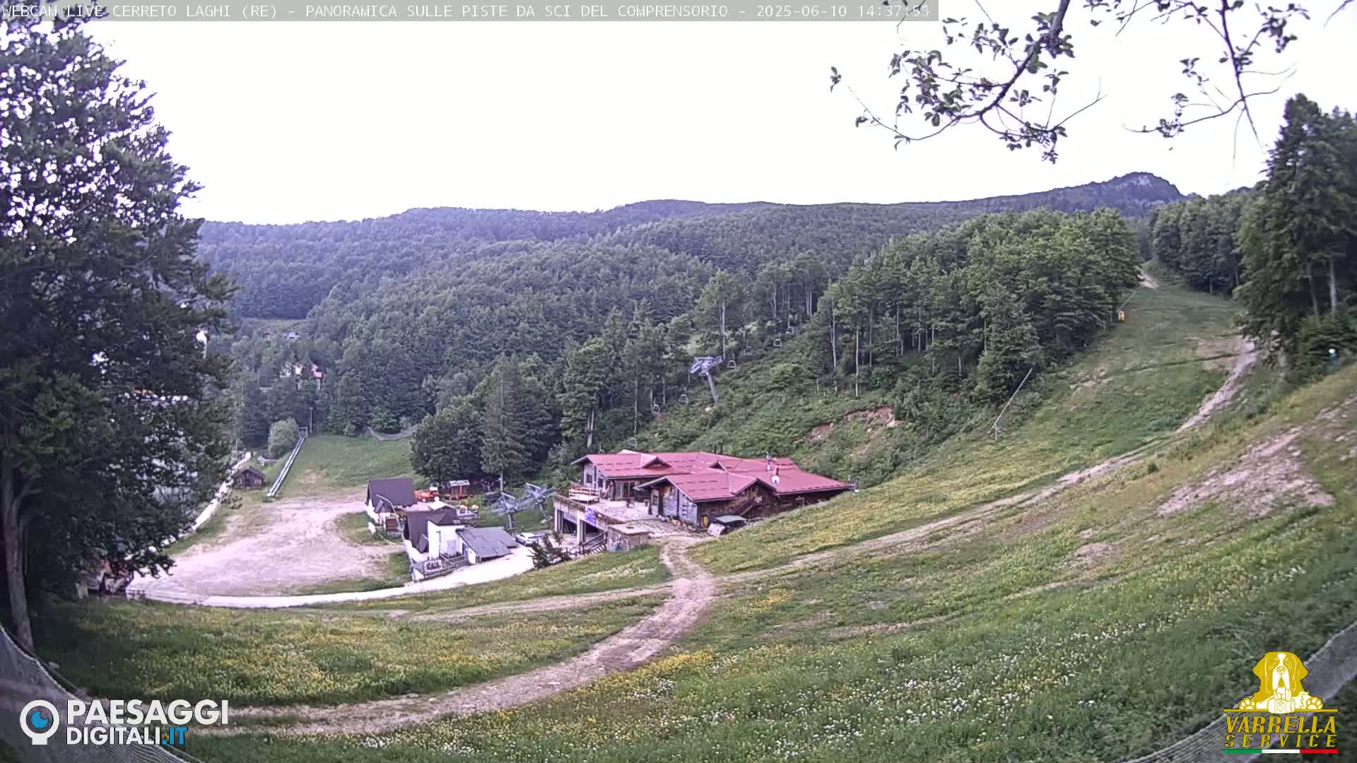 A mostly sunny view of a ski resort in the summer, showing grassy slopes, a few buildings, and a chairlift.