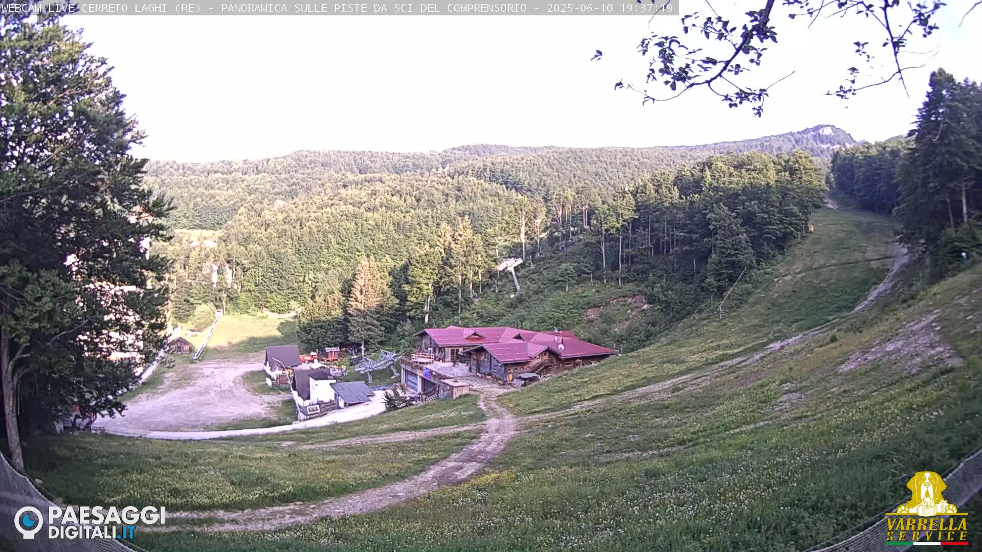 A mostly sunny view of a ski slope in the summer, showing a grassy hill with a building at the bottom and forested hills in the background.