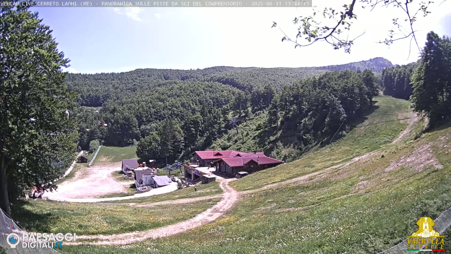 A mostly sunny, high-angle view of a grassy hillside with a ski resort building and empty ski slopes.