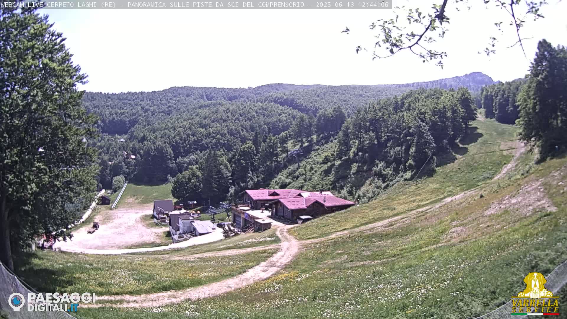 A high-angle view of a grassy hillside with a ski resort building and several smaller structures, under sunny skies.