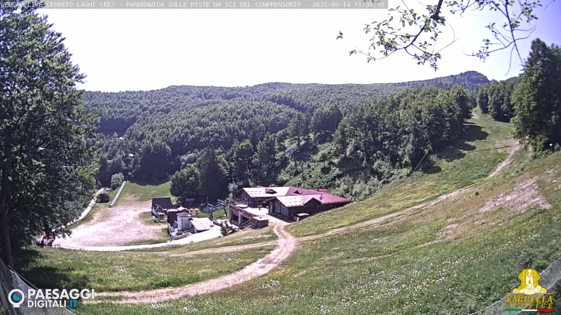 A grassy hillside slopes down to a building complex nestled among green trees under a mostly sunny sky.