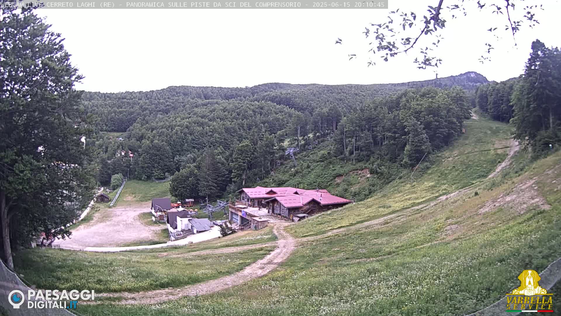 A mostly clear day reveals a ski resort in a mountain valley, with green grass covering the ski slopes and a reddish-brown building near the bottom.