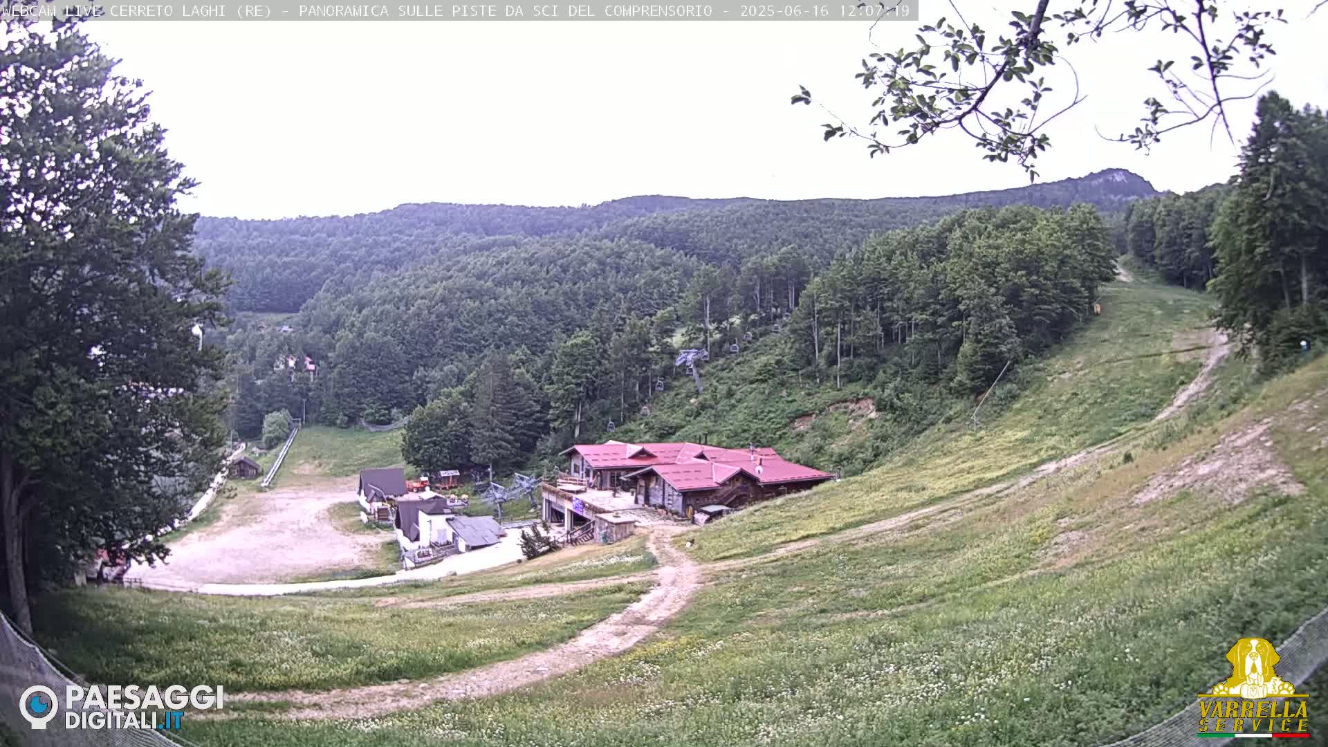 A mountain ski resort with a red-roofed building is shown, surrounded by green hills and a mostly clear sky.