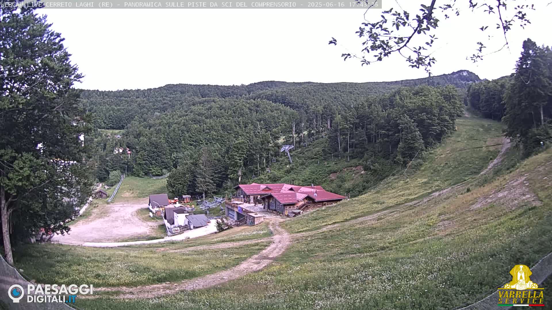 A mostly clear day reveals a ski resort in the mountains; a large building sits near the base of a grassy ski slope, with more buildings and a chairlift visible further up the hill.