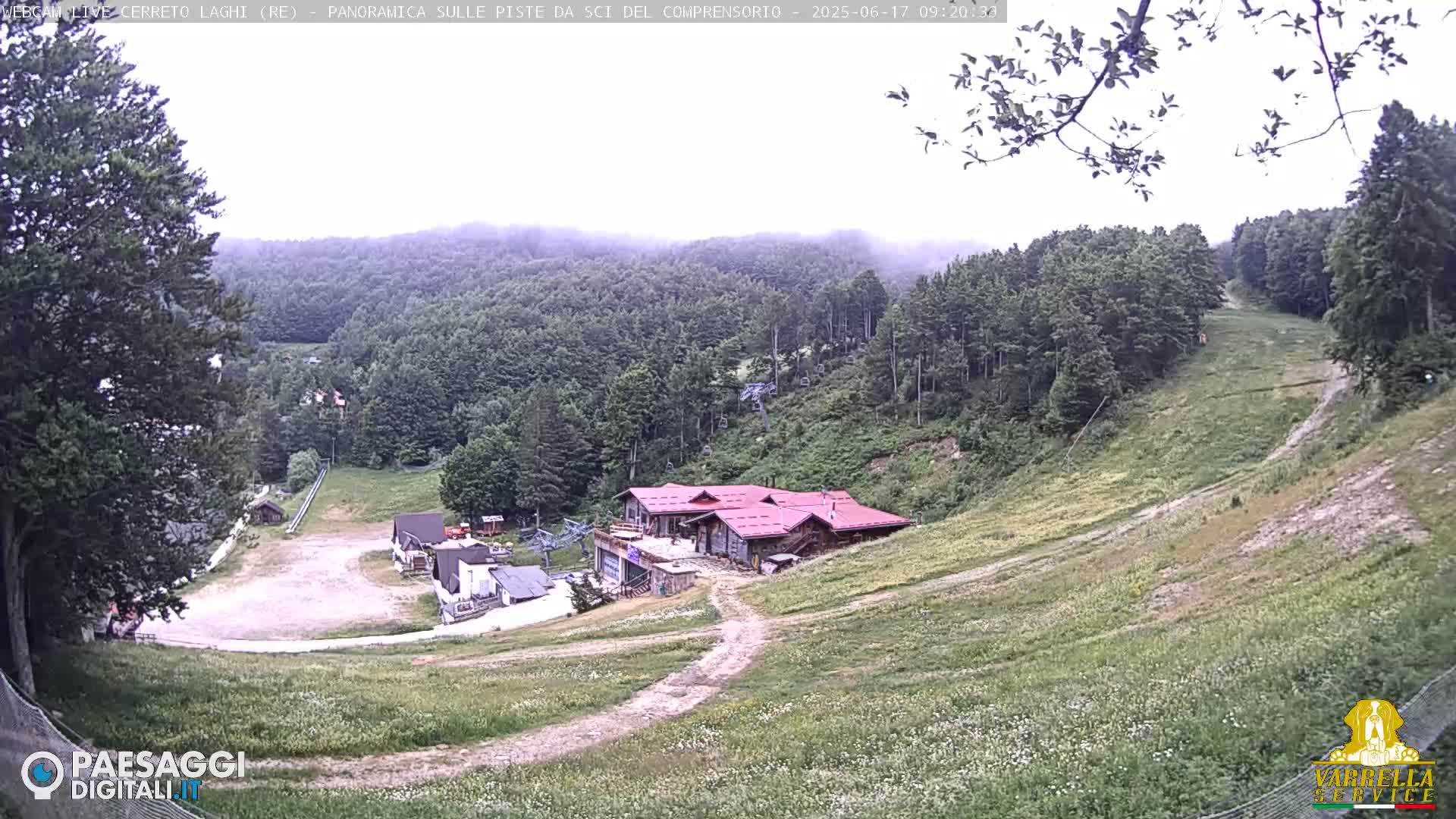A mostly cloudy view of a ski resort in the summer, showing grassy ski slopes, a main lodge, and surrounding green hills.