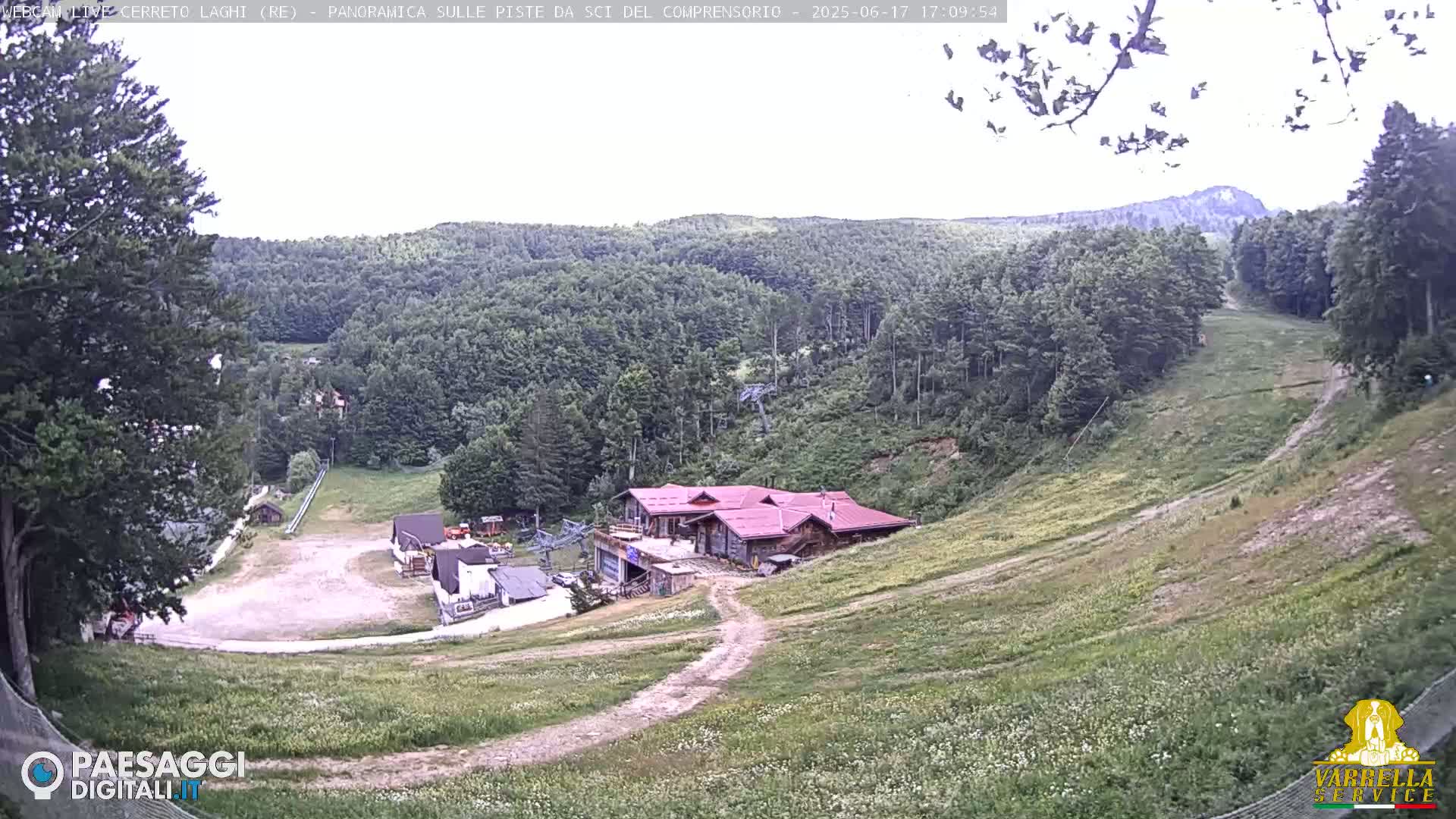 A mostly clear day overlooking a ski slope with sparse vegetation, a red-roofed building, and a wooded hillside in the background.
