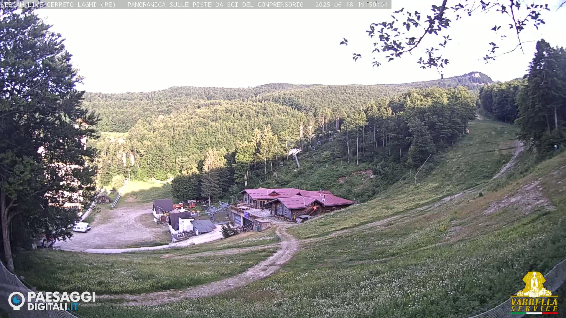 A mountainside ski resort, mostly grassy with some trees and a building, is visible under a mostly clear sky.