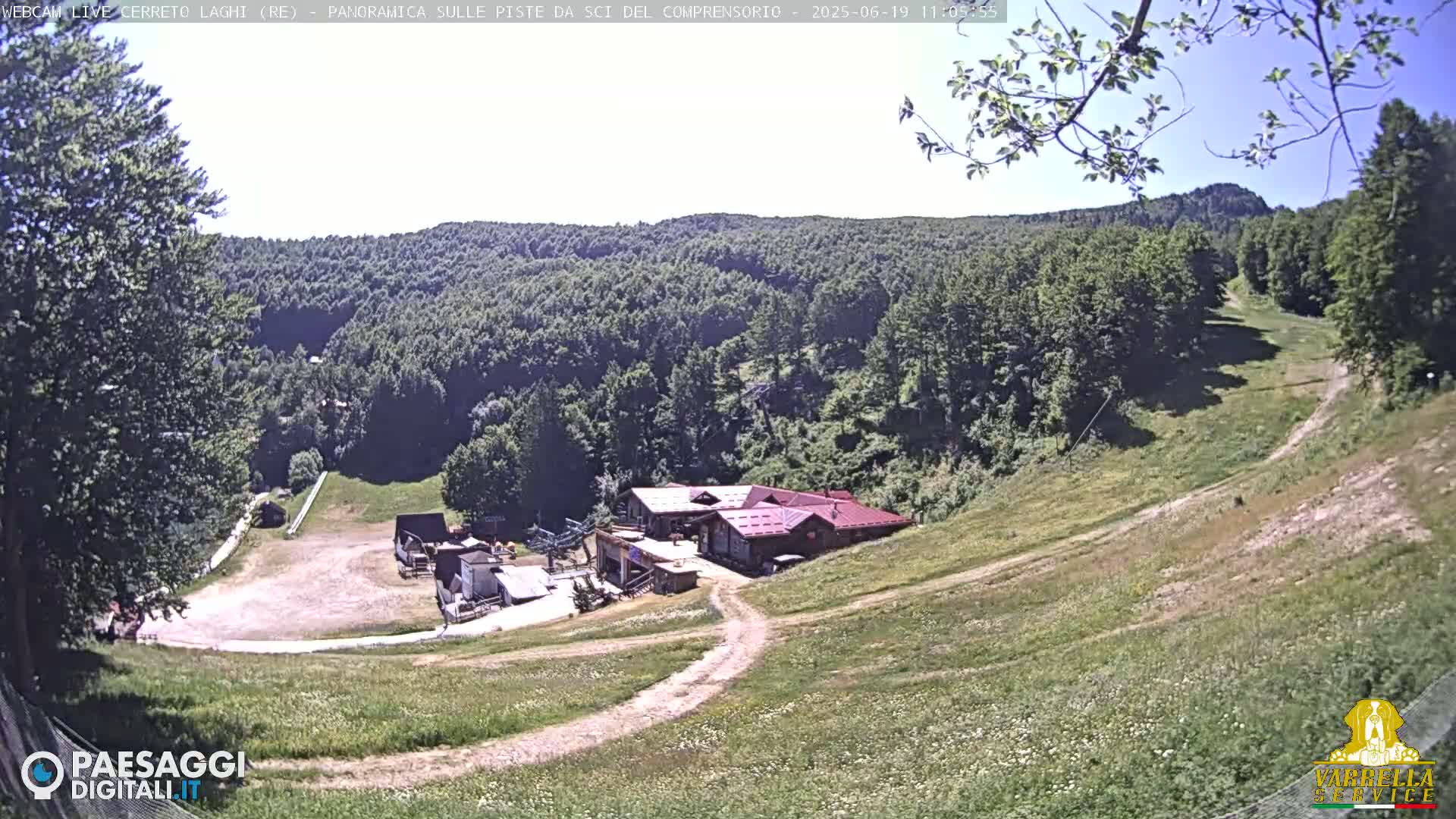 A high-angle view on a sunny day shows a grassy ski slope leading down to a building complex nestled among green, forested hills.