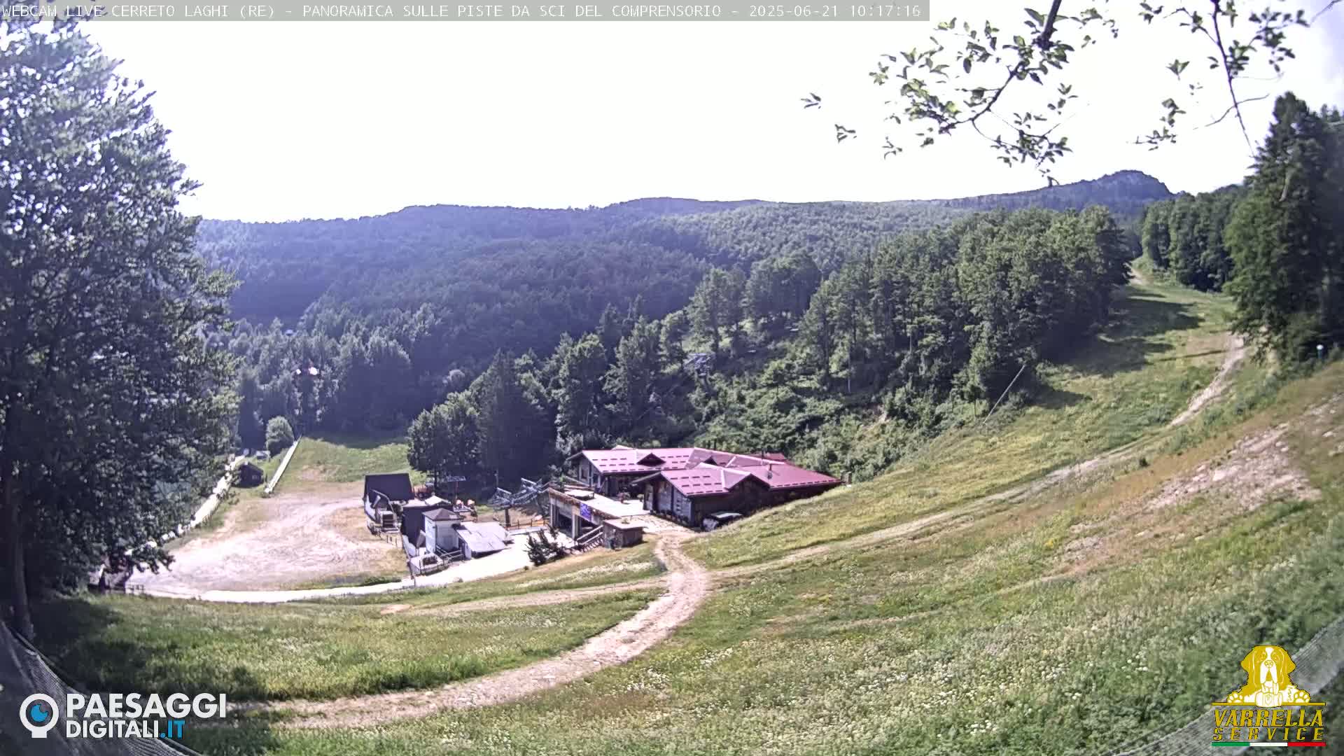 A grassy hillside slopes down toward a ski lodge and empty ski runs, under a bright, sunny sky.