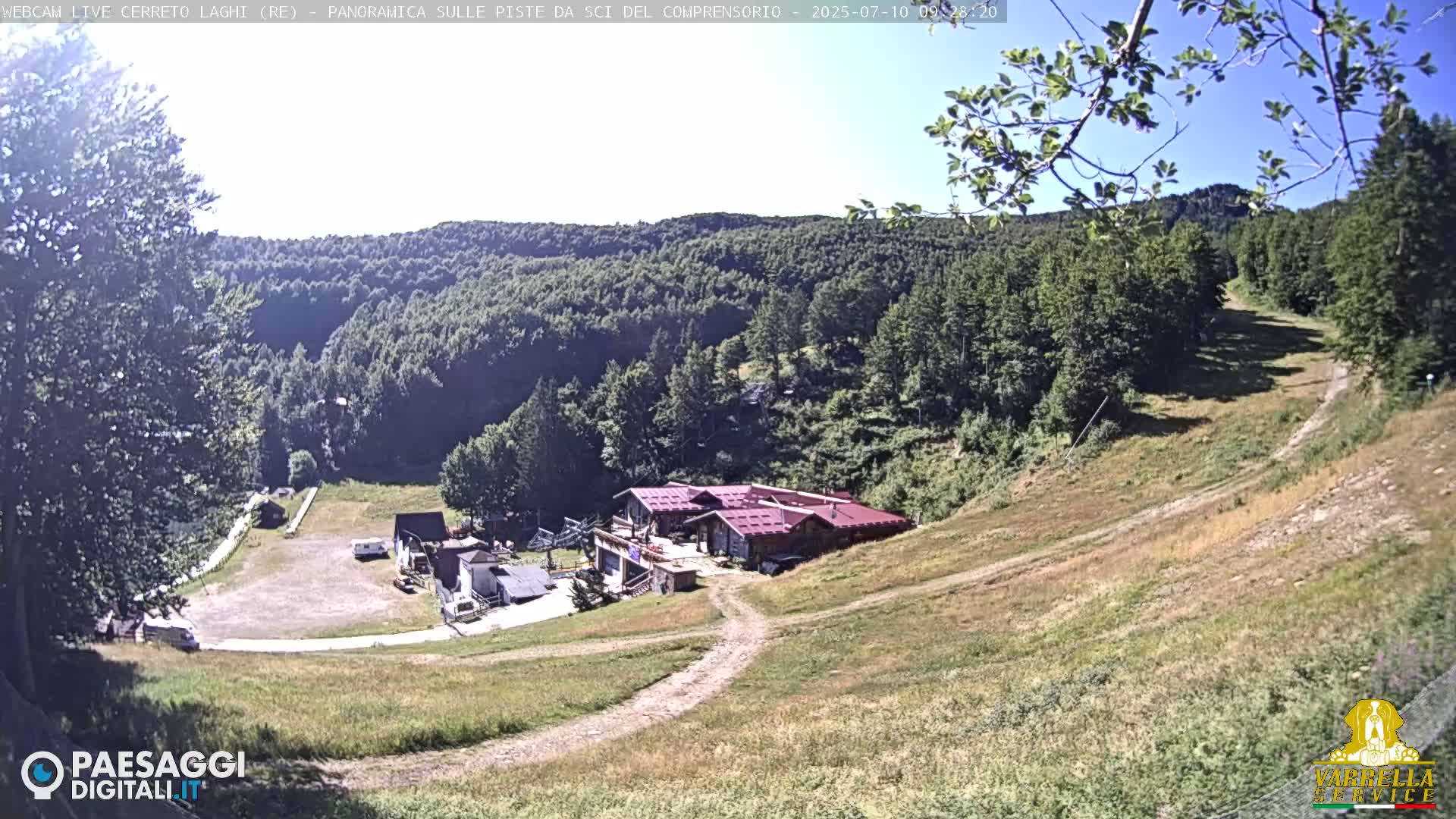 A sunny view of a mountainside ski resort with a building, parking area, and grassy slopes.