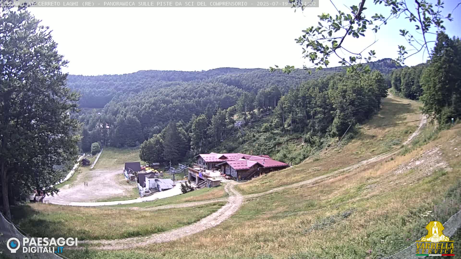 A ski resort in a hilly, forested area on a sunny day shows a building at the bottom of a grassy, dirt slope leading up to a mountain.