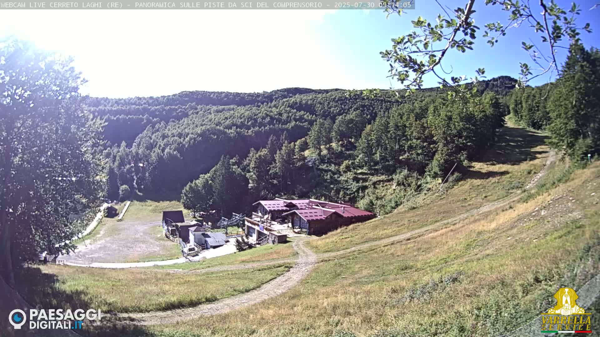 A high-angle view of a mountainside building complex with surrounding grassy slopes and a forest in the background under sunny conditions.