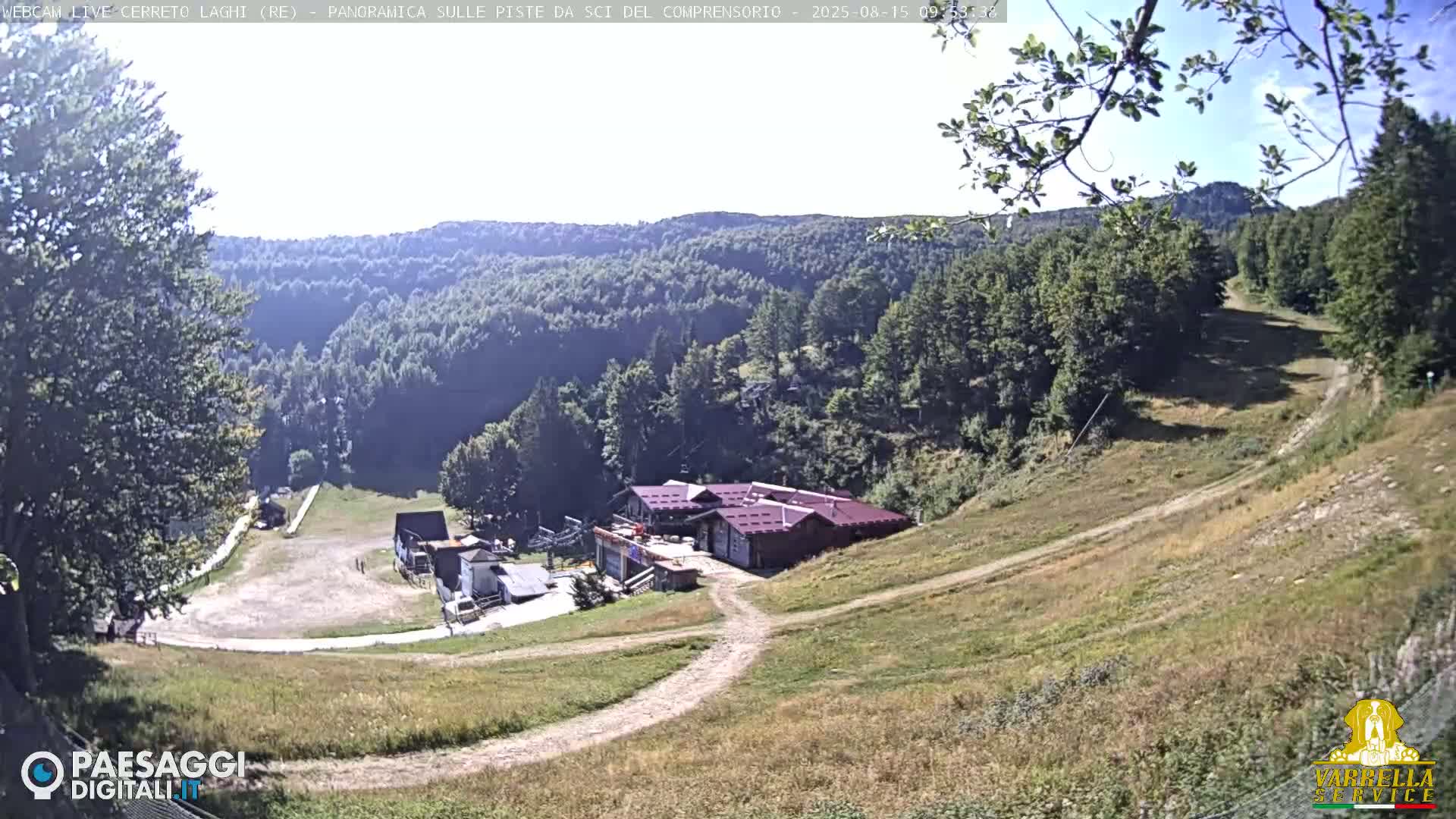 A ski resort in the mountains on a sunny day shows a grassy hillside leading to a building and ski lift equipment.