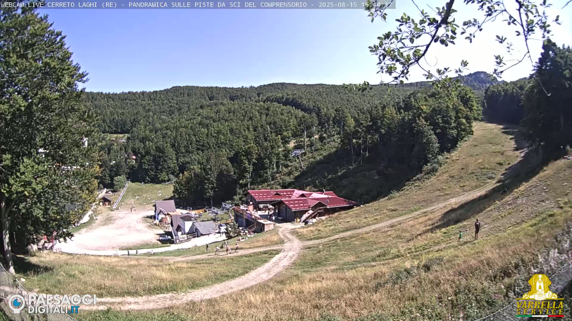 A ski resort in the mountains on a sunny day shows a grassy hillside leading to a building and ski lift equipment.
