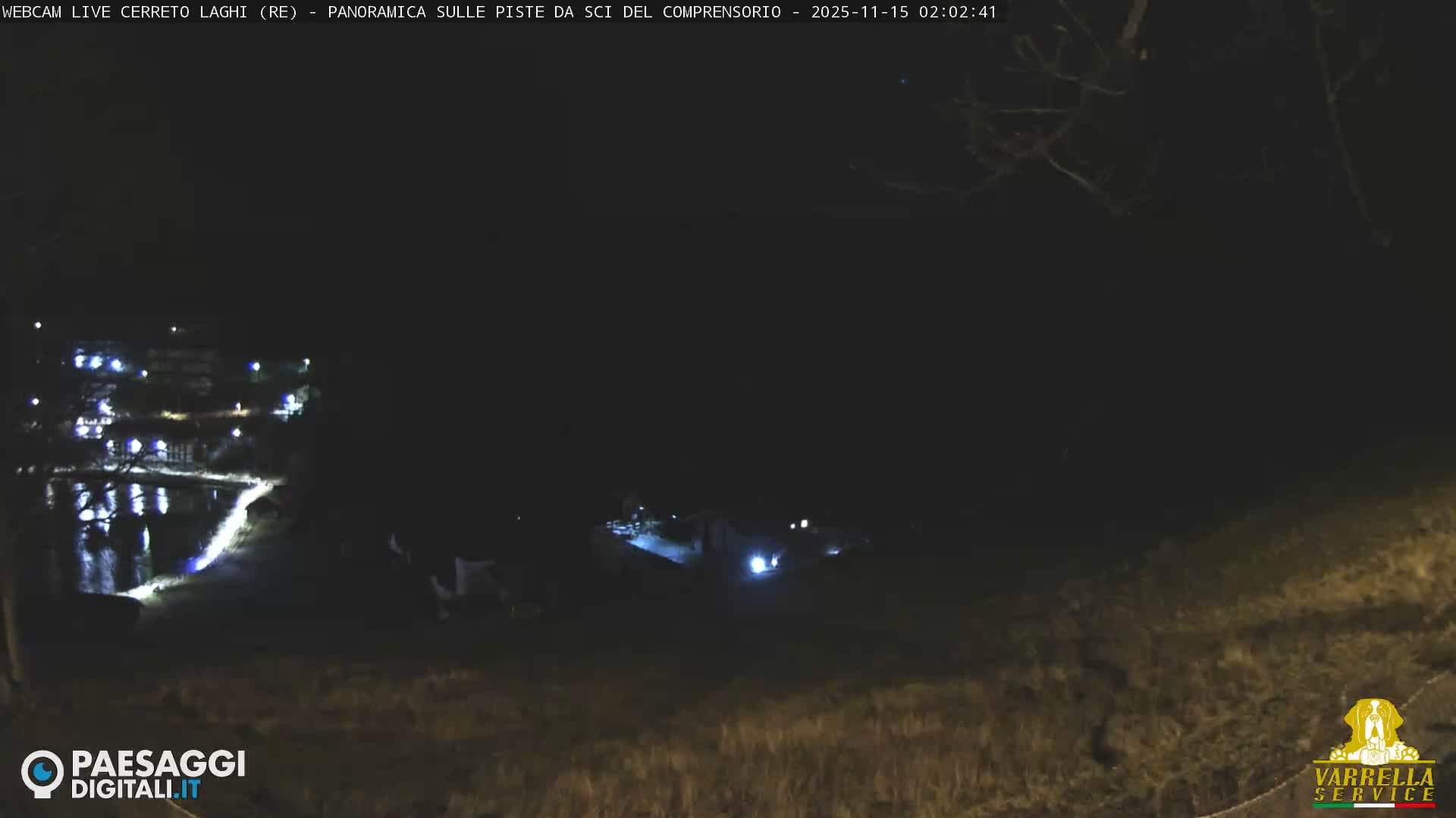 A clear, dark night reveals a tranquil scene of a village with illuminated buildings and a brightly lit path reflecting on a calm body of water, with faint vehicle lights visible in the distance.
