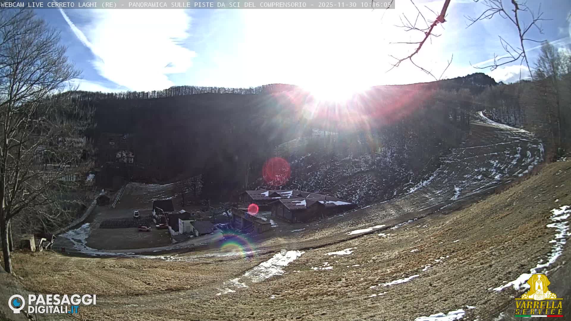 Under sunny, clear conditions, a snow-dusted mountain landscape features a cluster of buildings nestled amidst brown grassy slopes and bare trees.