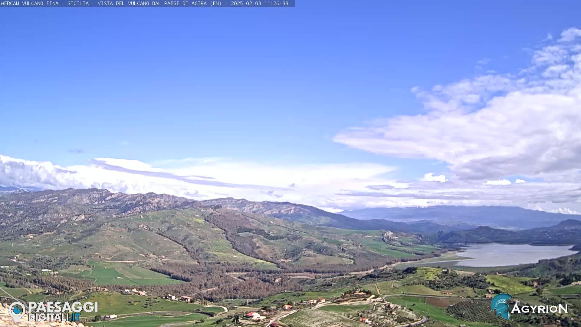 A mostly clear, blue sky with scattered clouds overlooks a sprawling landscape of rolling green hills, farmland, and a lake in the distance.
