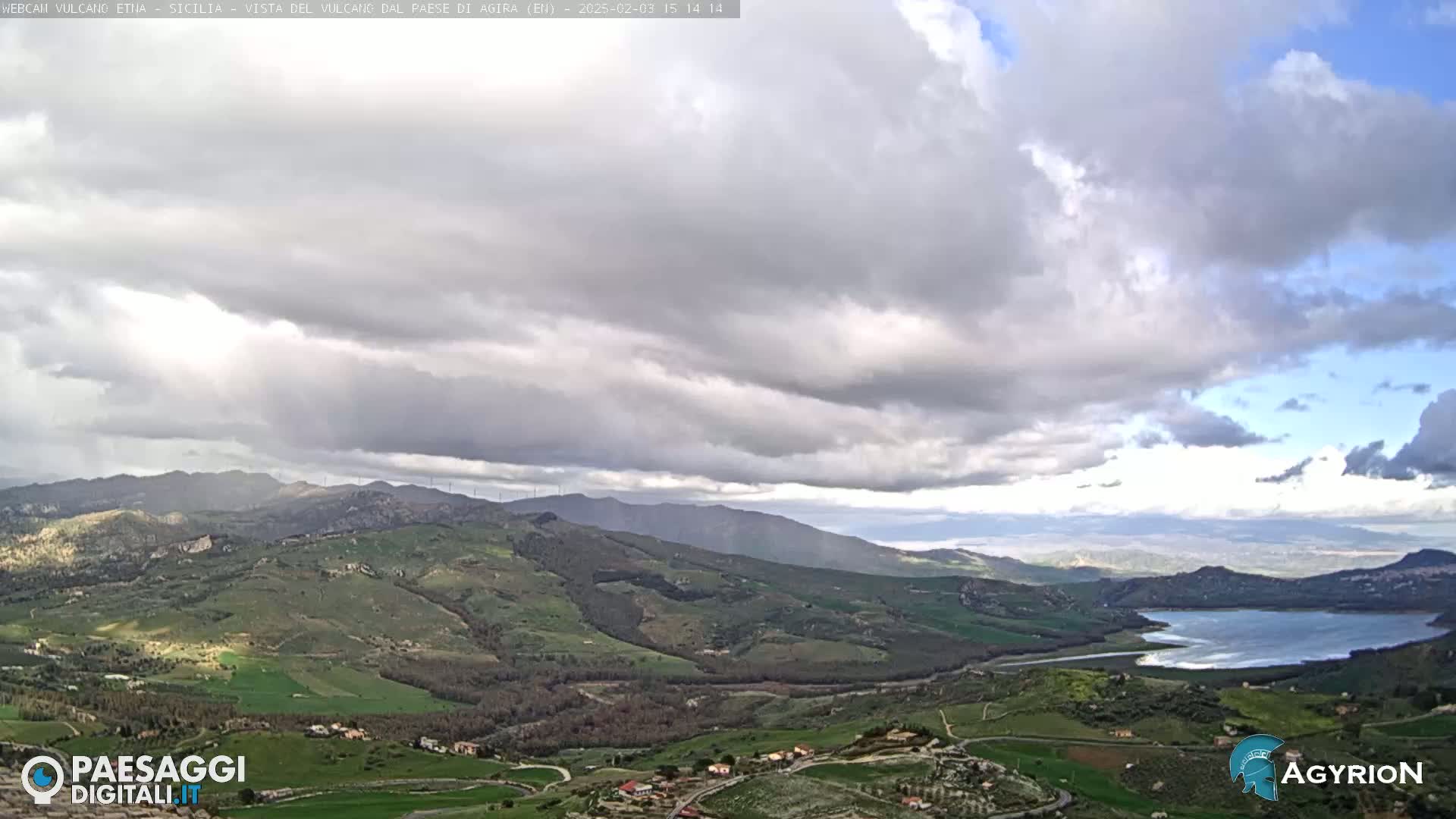 A vast, hilly landscape under a mostly cloudy sky features a body of water in the distance.