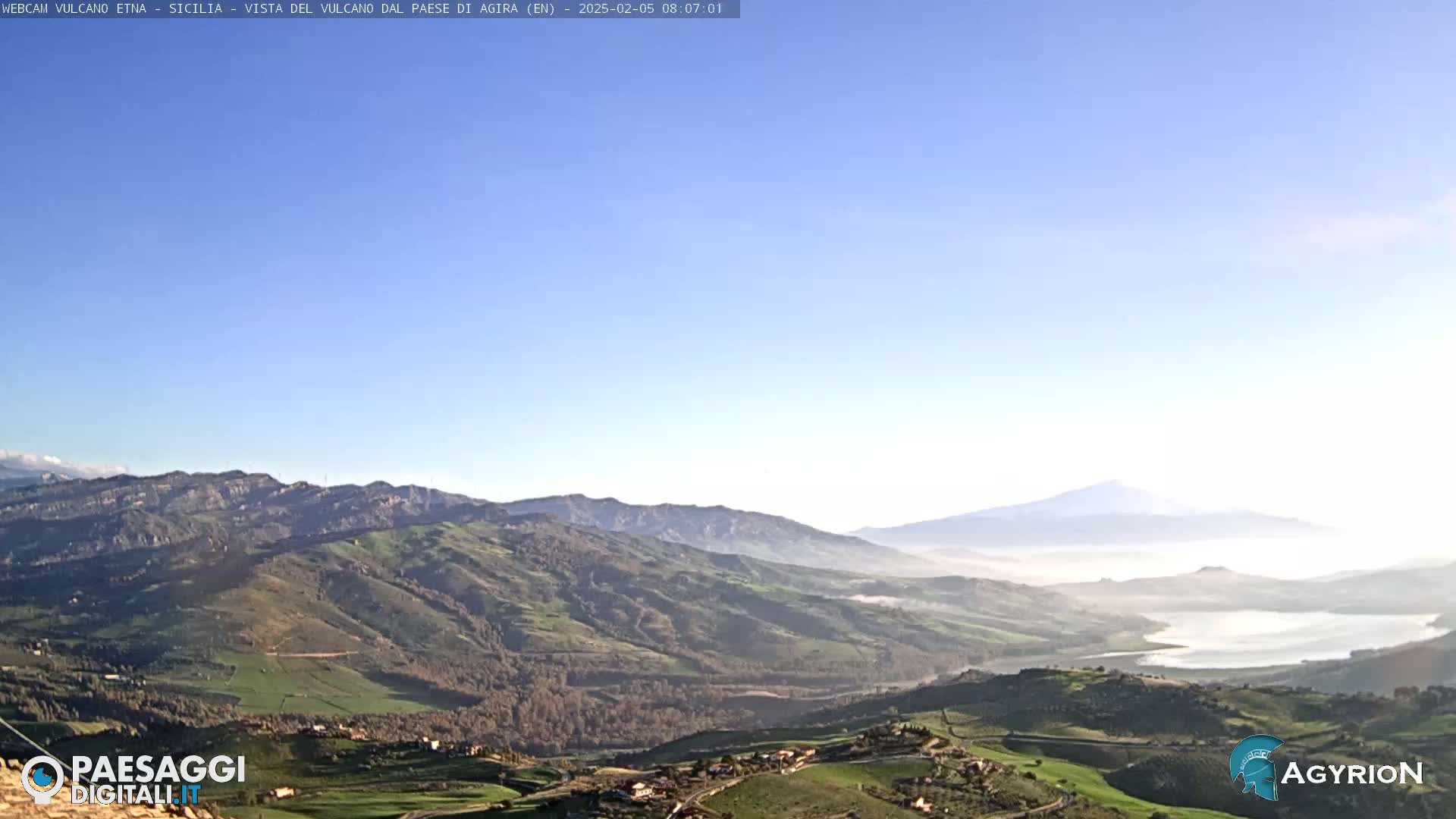 A distant, snow-capped volcano is visible over a valley of rolling green hills and a lake under a clear blue sky.