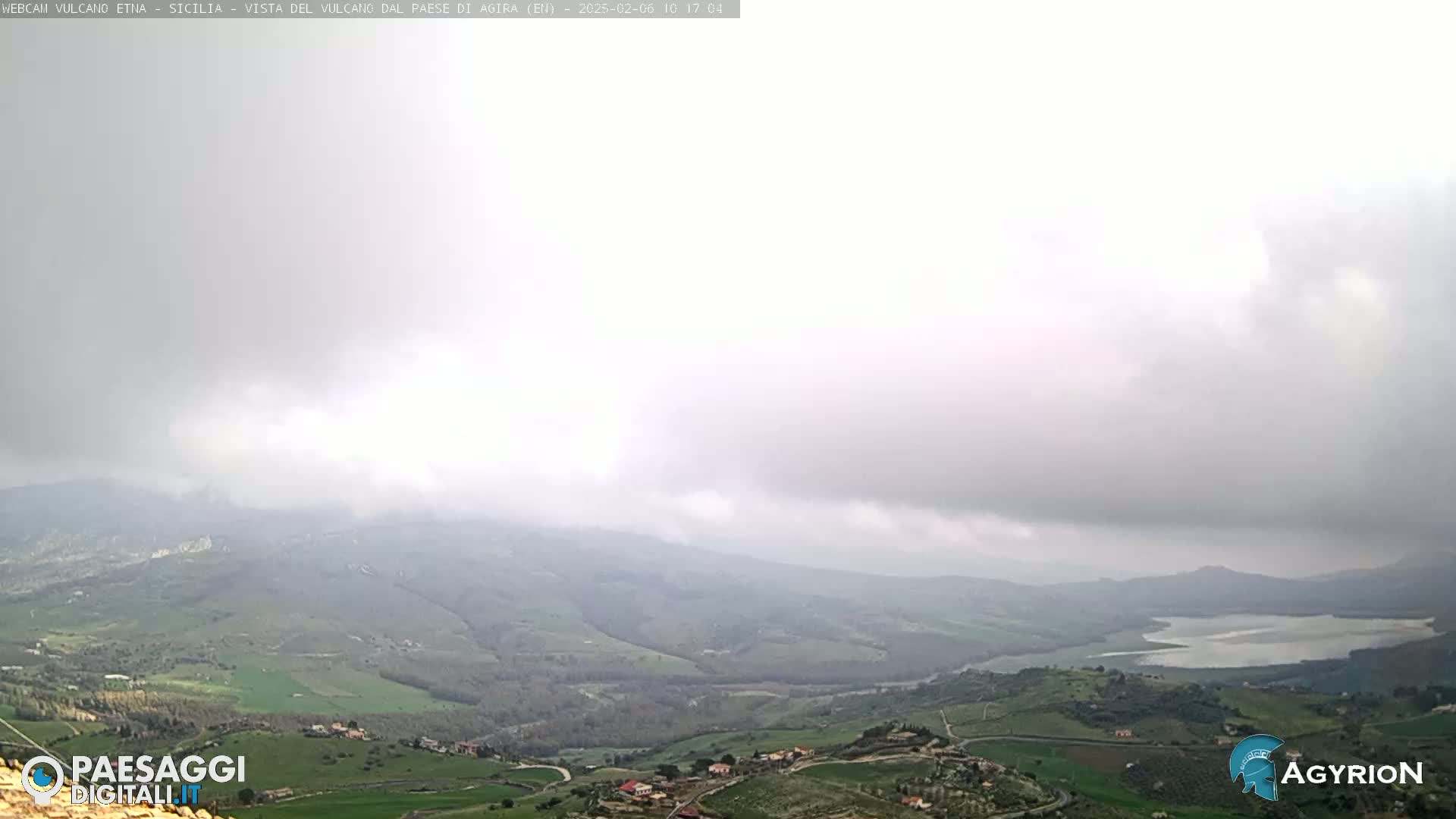 A hazy, overcast sky hangs over a verdant valley dotted with small settlements and a large body of water in the distance.
