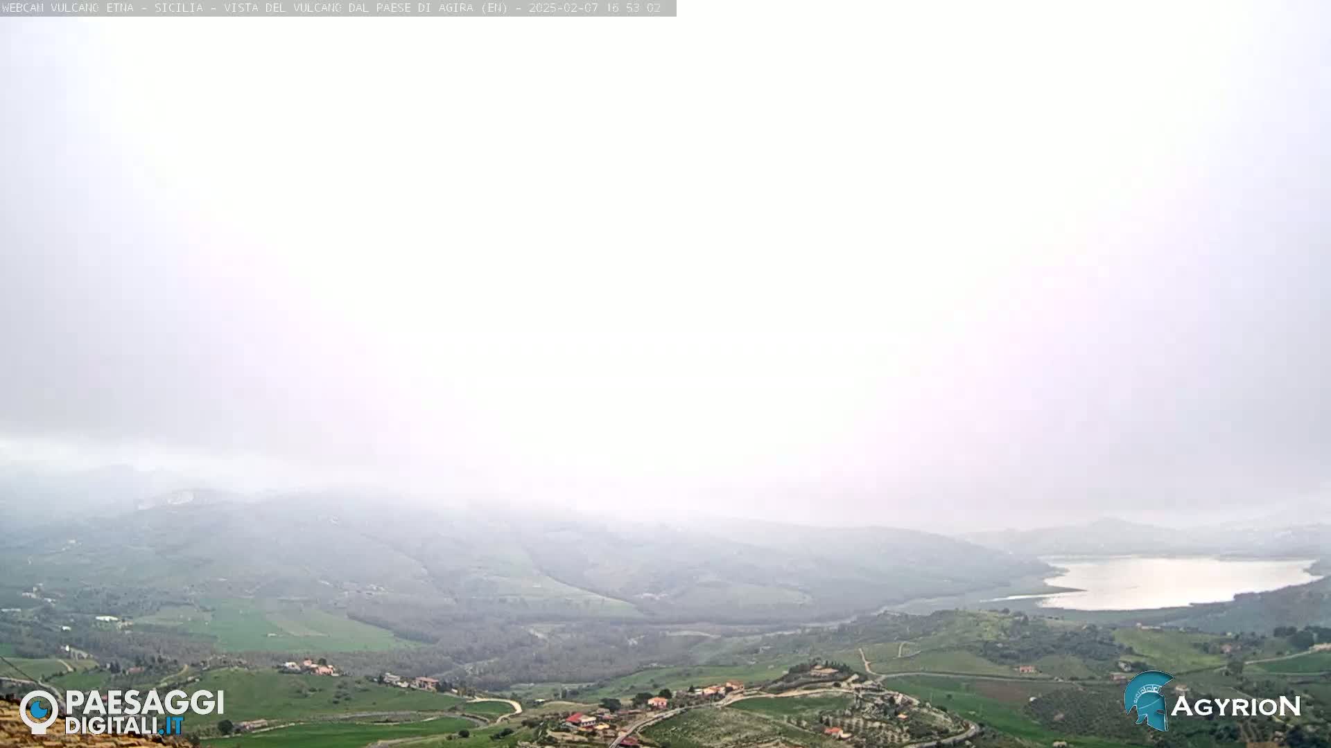 A hazy, overcast view of a valley with a lake and scattered settlements, stretching to rolling hills in the distance.