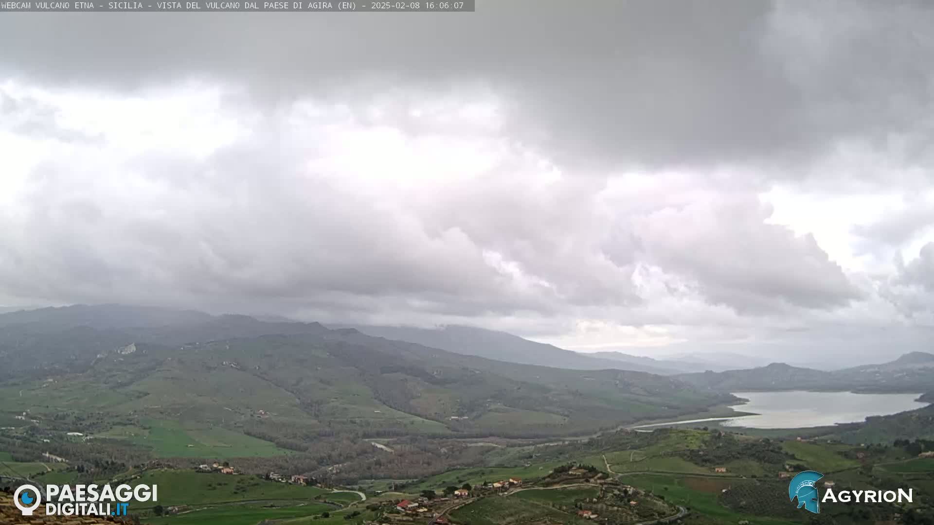 A sprawling valley with rolling green hills and a lake is visible under a mostly cloudy sky.