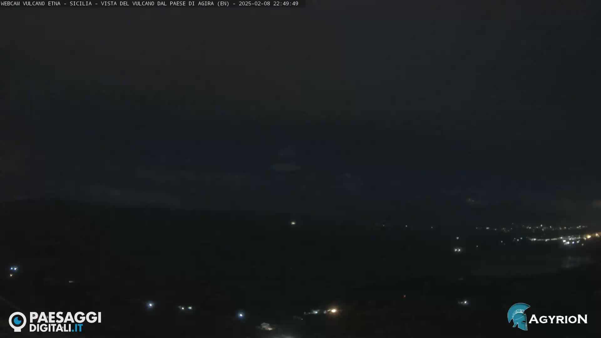 A nighttime long shot shows a dark, mostly obscured landscape with a few scattered lights visible in the distance under a dark sky.