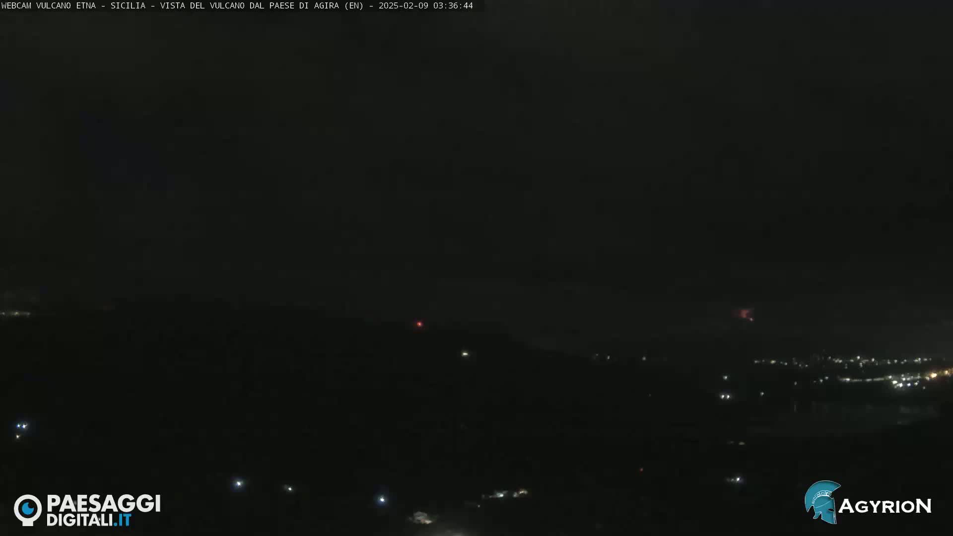 A nighttime long shot shows a dark landscape with a few scattered lights, and faint reddish glows possibly indicating volcanic activity in the distance under a dark, clear sky.