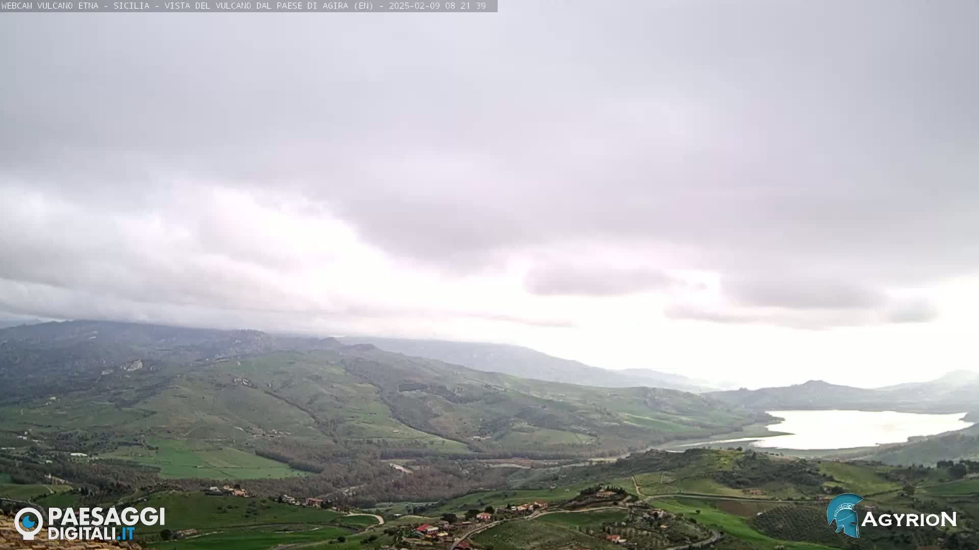 A panoramic view of a valley and a lake under a mostly cloudy sky.
