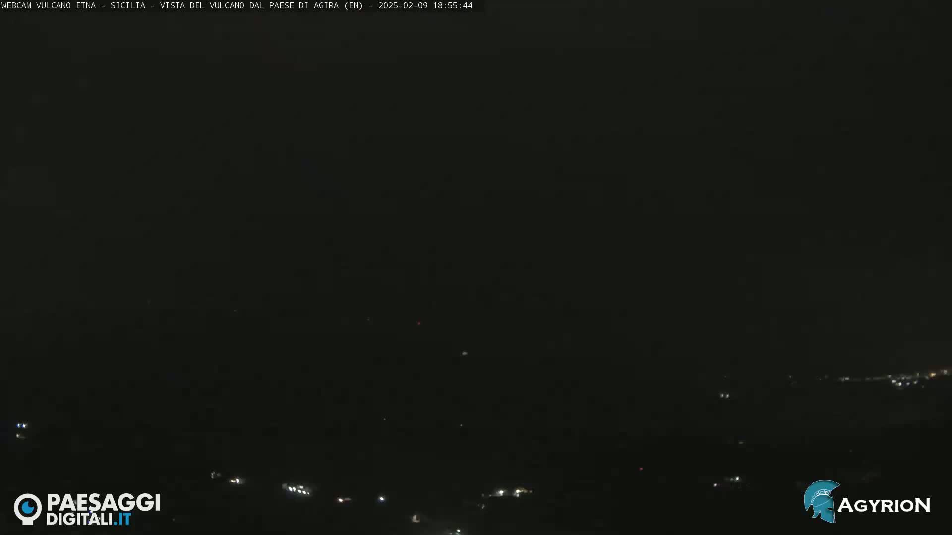 A nighttime long shot of a dark landscape with a few distant lights.