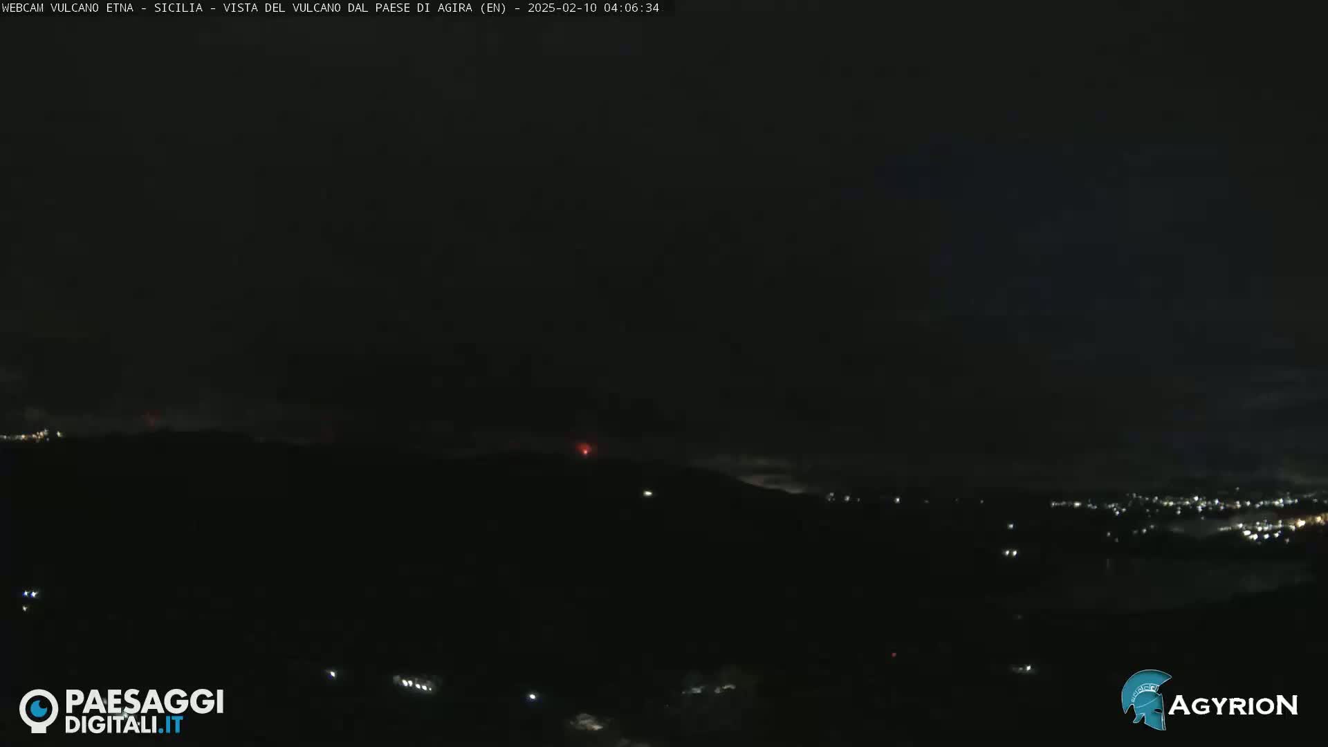 A nighttime long shot of a volcano erupting with a faint red glow, viewed from a distance over a dark landscape with sparsely lit areas.