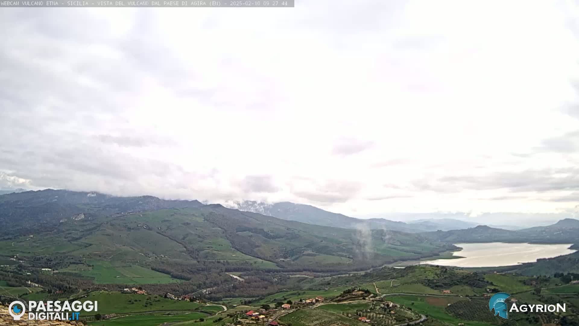 A sprawling, verdant landscape unfolds under a mostly cloudy sky, featuring rolling hills, a tranquil lake, and a distant mountain range partially shrouded in mist.