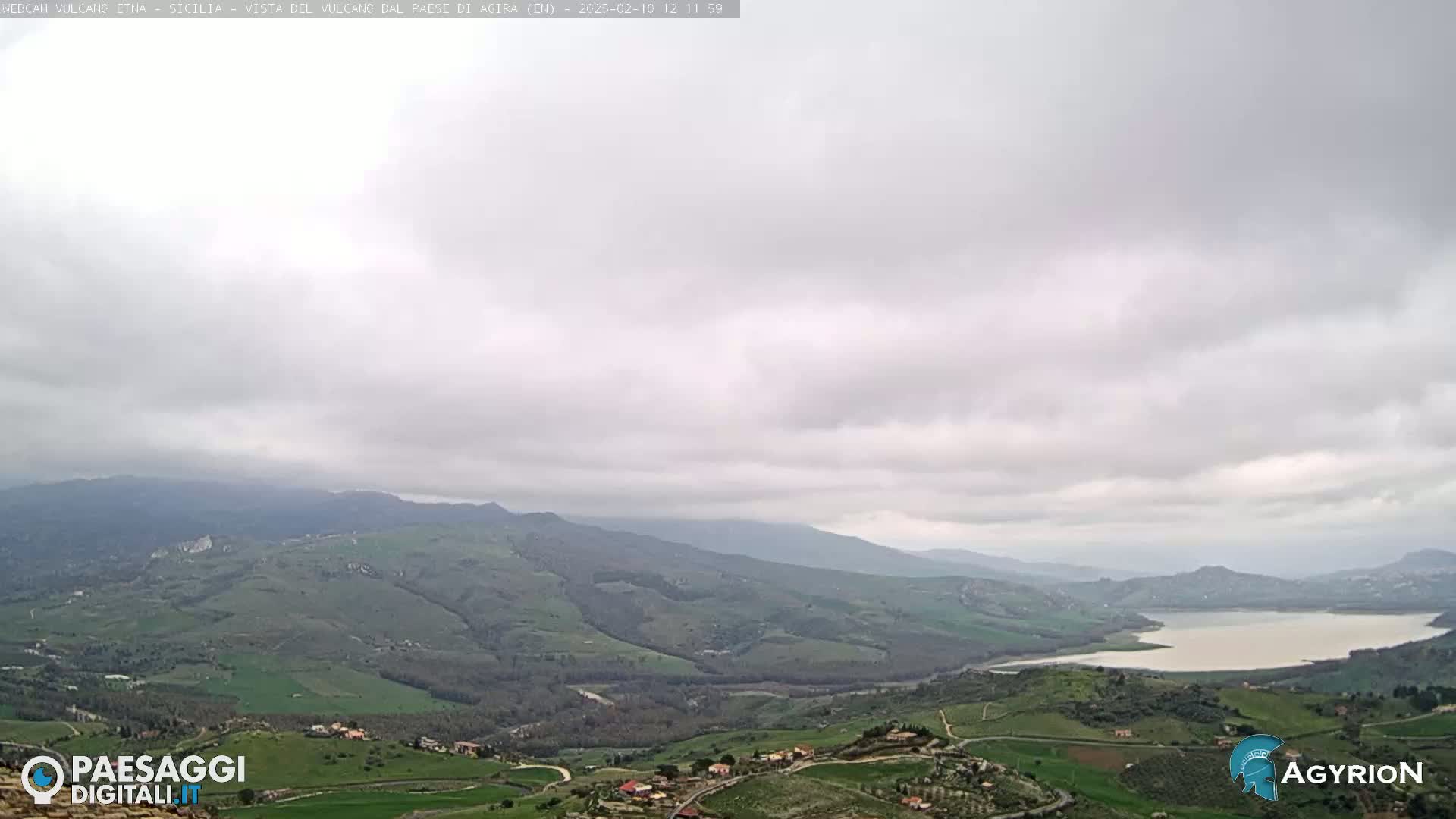A wide, overcast view of a valley with rolling green hills, a lake, and distant mountains.