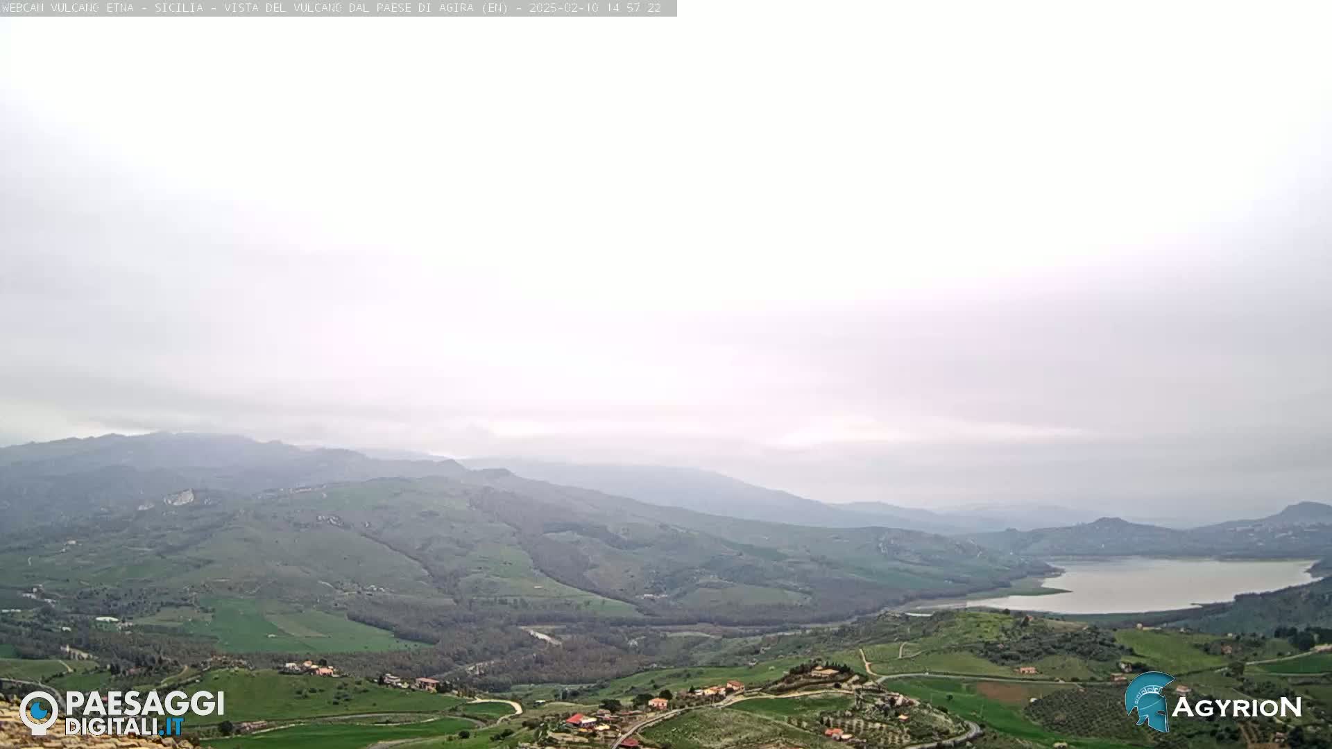 A hazy, overcast day reveals a sprawling landscape of rolling green hills, a calm lake, and a small village nestled in a valley.
