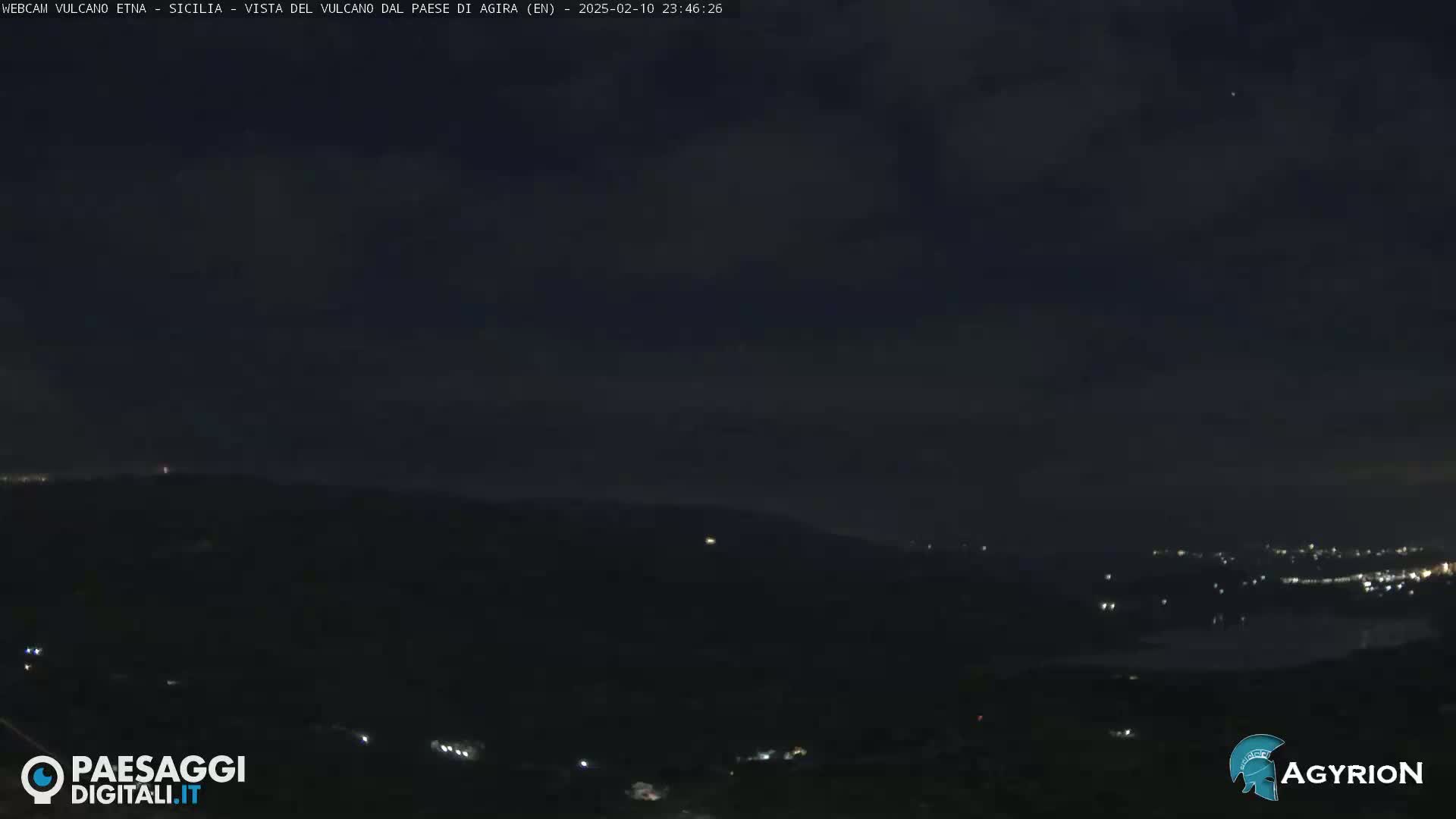A nighttime view of dark hills and a distant town illuminated by lights under a mostly cloudy sky.