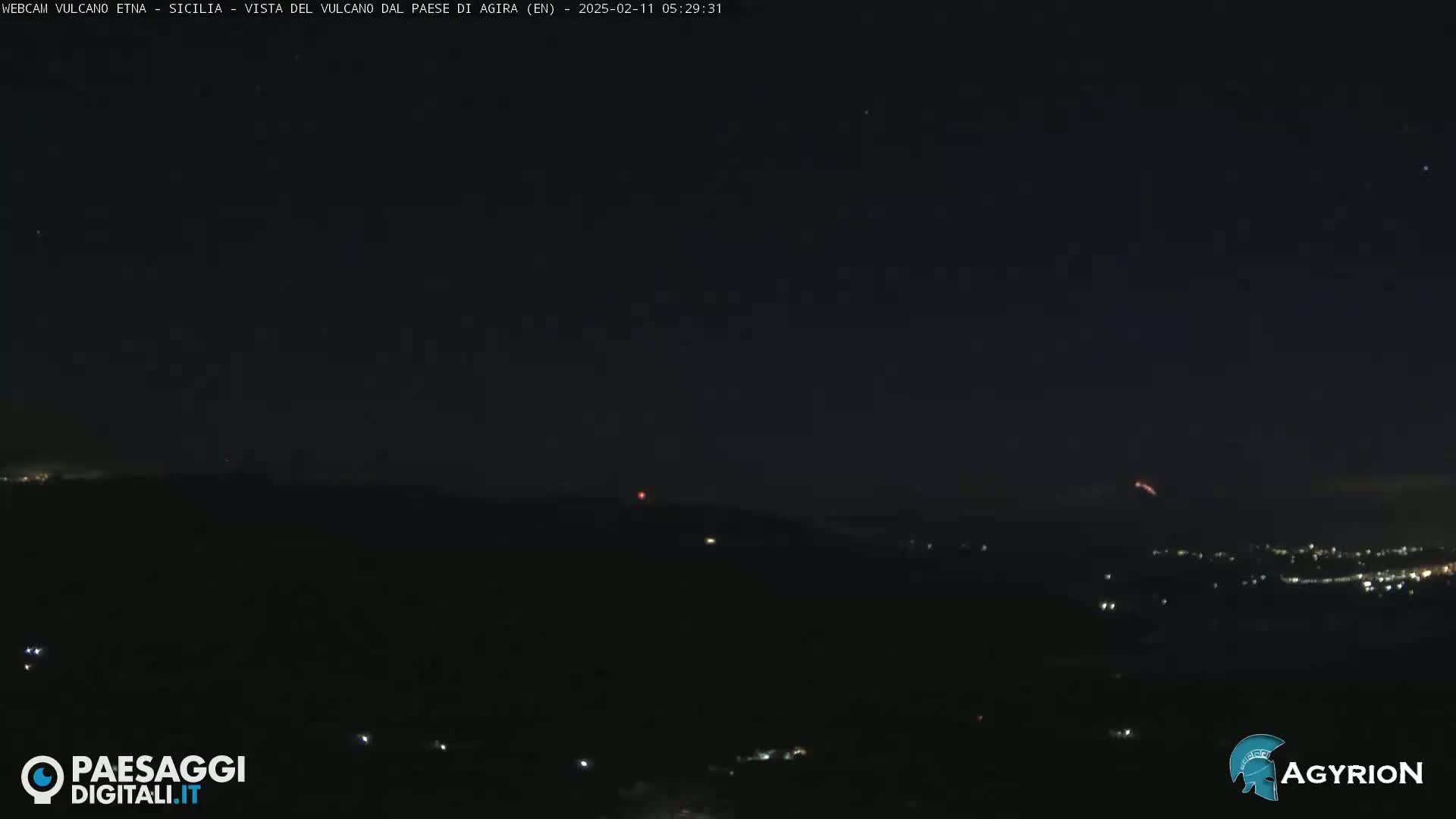A nighttime long shot of a dark landscape with a few distant lights and a faint reddish glow in the sky.
