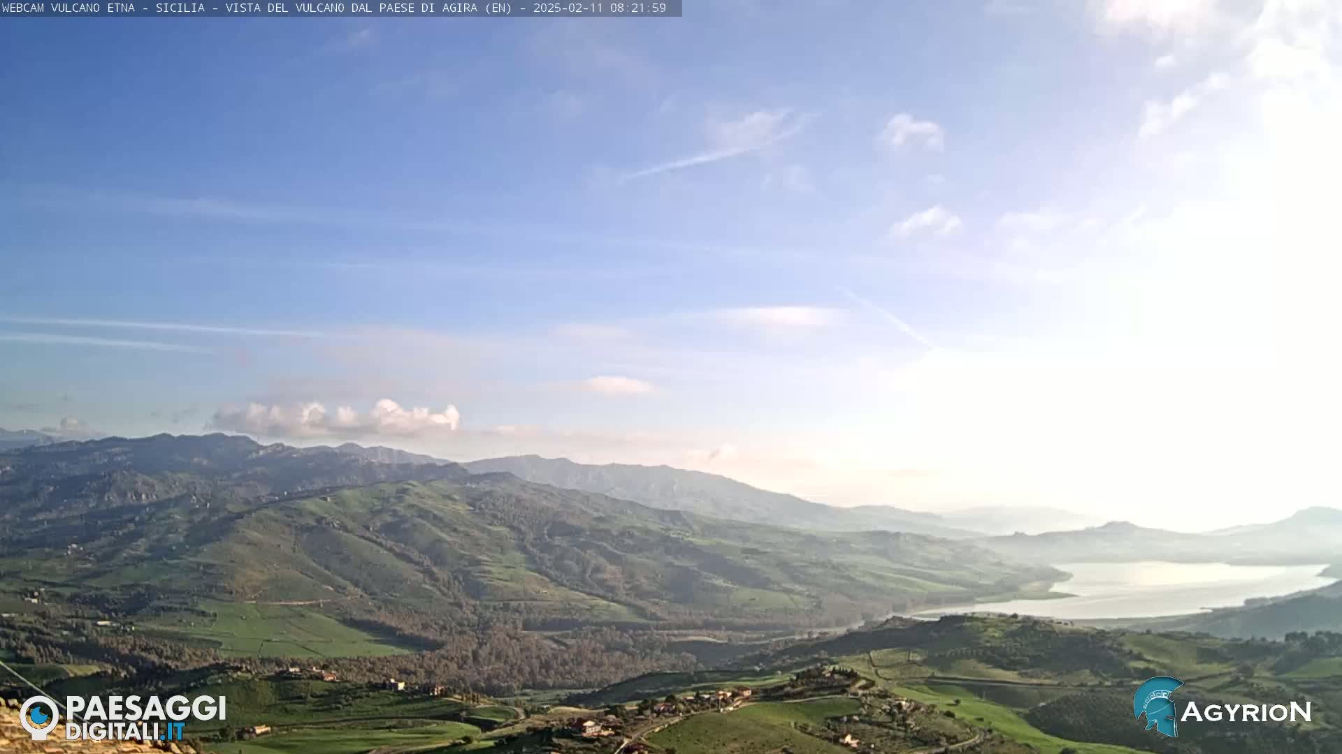 A vast, rolling landscape of green hills and valleys stretches to a distant lake under a mostly clear, light blue sky with a few scattered clouds.