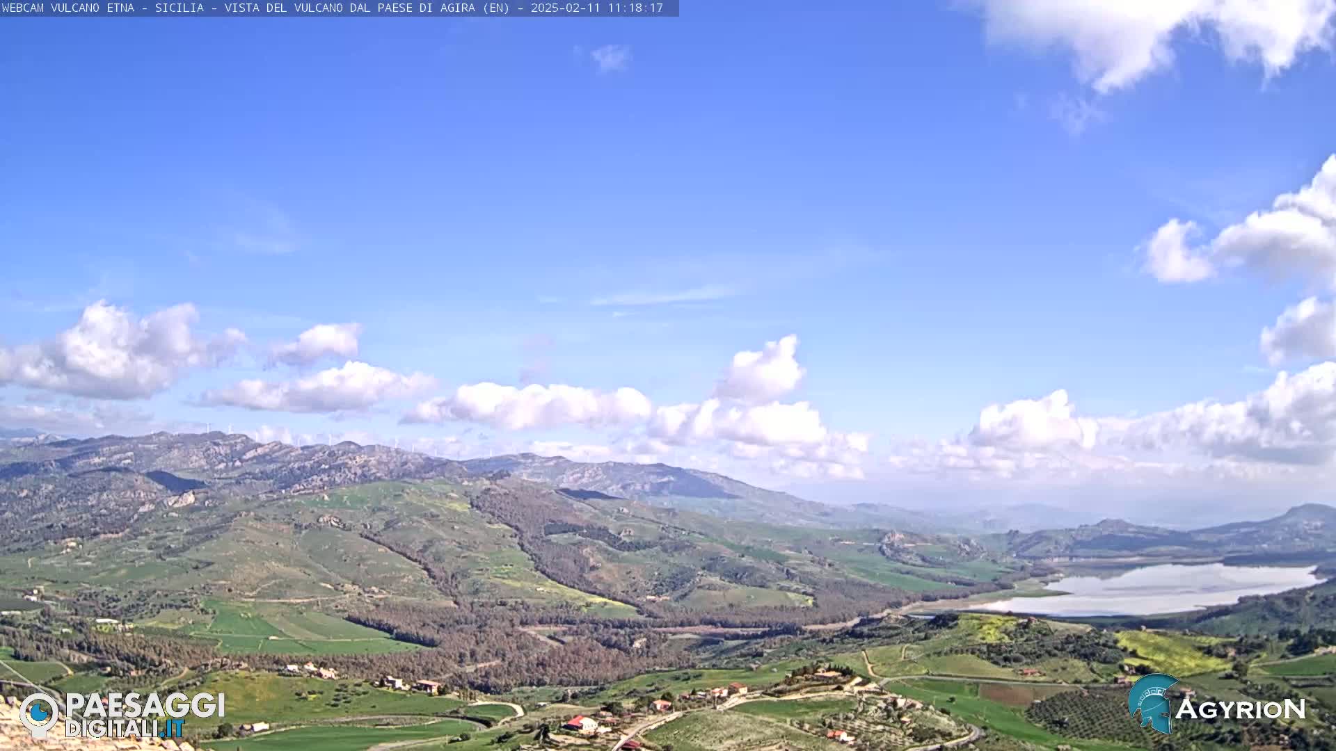A vast, hilly landscape under a mostly clear blue sky with scattered cumulus clouds features a large body of water in the distance.