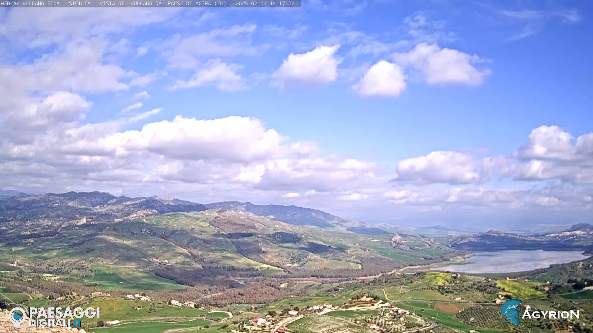 A partly cloudy sky overlooks a sprawling valley of rolling hills and farmland, with a large body of water visible in the distance.