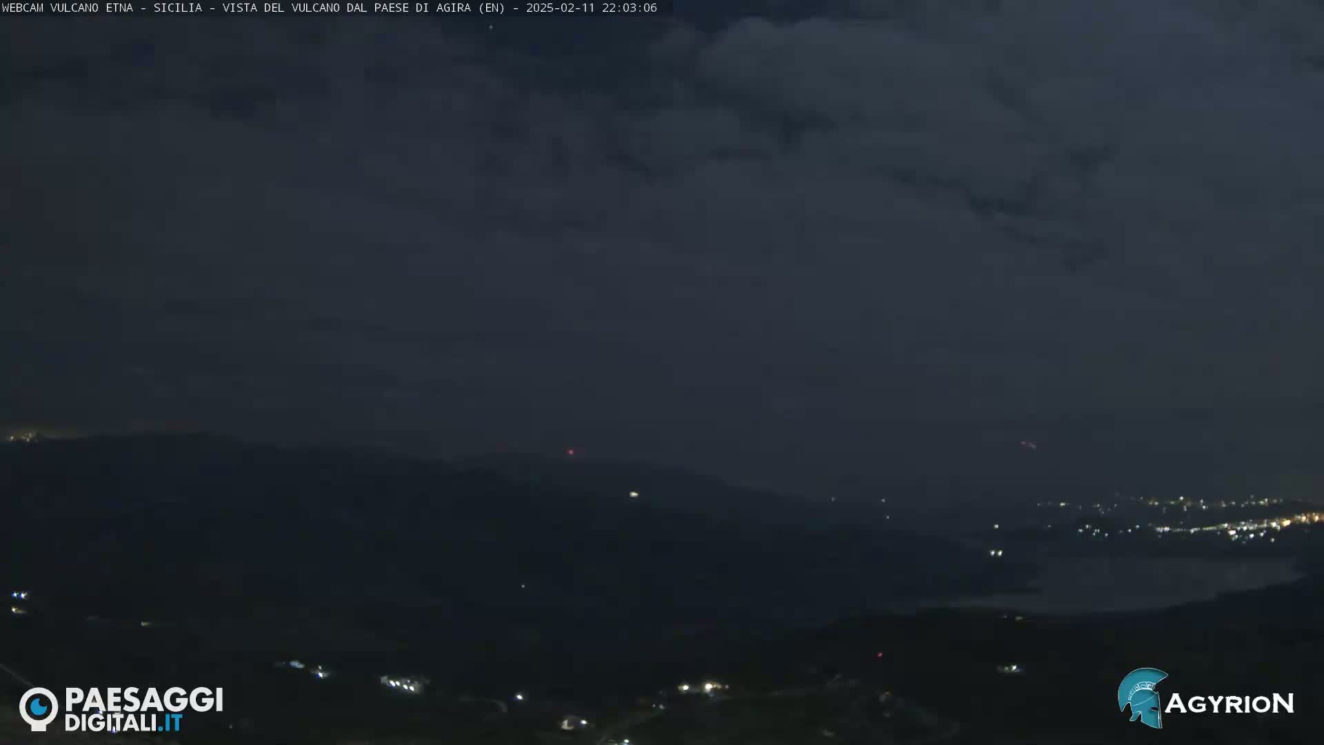A nighttime, cloudy view of dimly lit hills and a body of water in the distance.