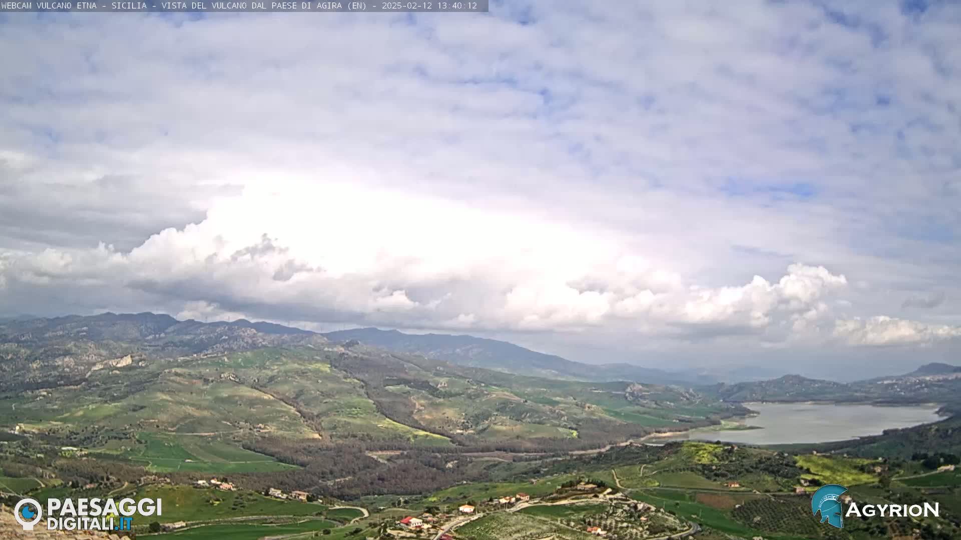 A partly cloudy day reveals a sweeping view of rolling green hills, farmland, and a lake nestled in a valley, with mountains in the distance.