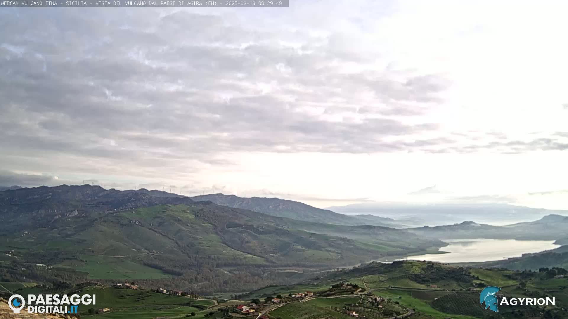 A hazy, overcast sky overlooks a vast, rolling landscape of green hills and valleys, with a lake visible in the distance.