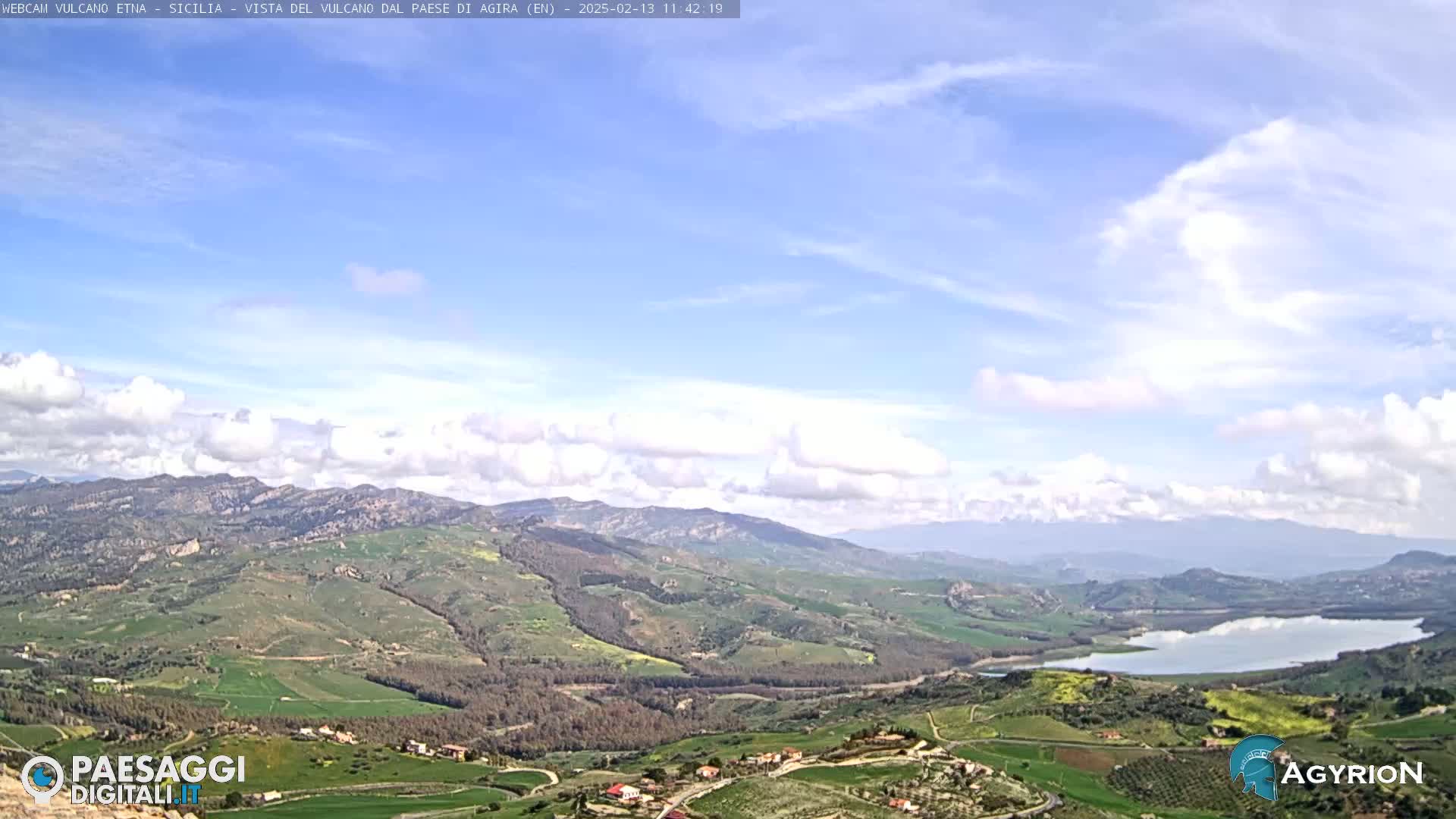 A partly cloudy sky overlooks a vast, verdant valley with a lake, rolling hills, and scattered farmlands.