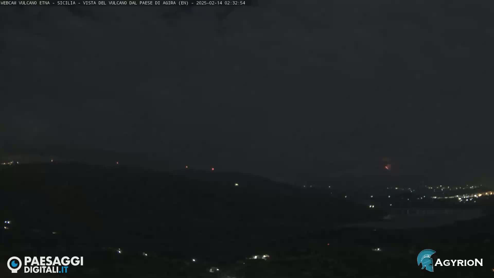 A nighttime, long shot of a dark, hilly landscape with a few faint lights and reddish glows in the distance under a mostly clear sky.