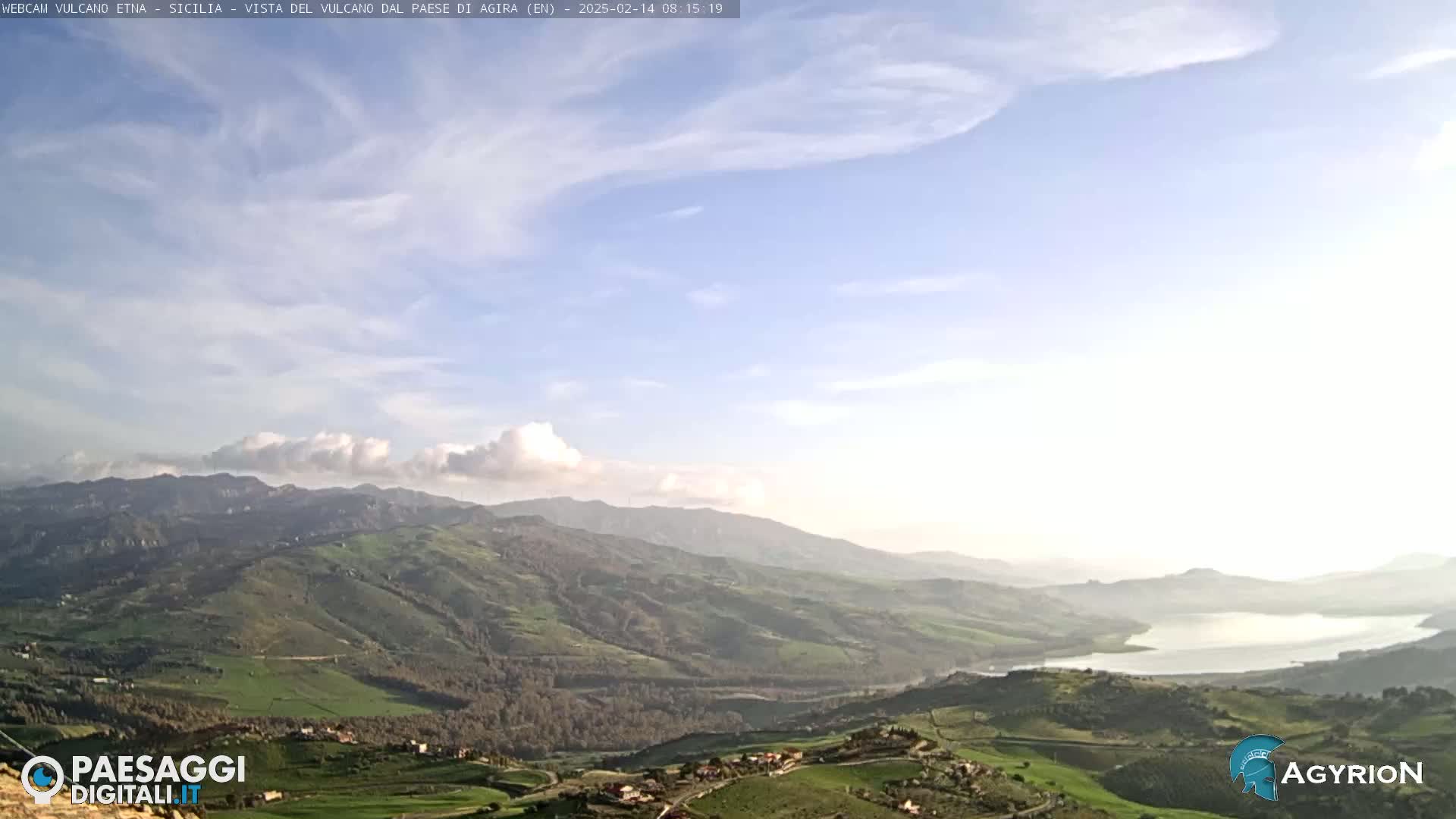 A partly cloudy sky overlooks a vast, rolling landscape of green hills and a lake in the distance.