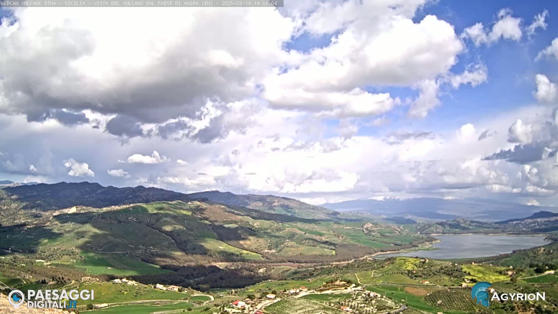 A partly cloudy sky overlooks a vast, hilly landscape dotted with villages and a large lake.
