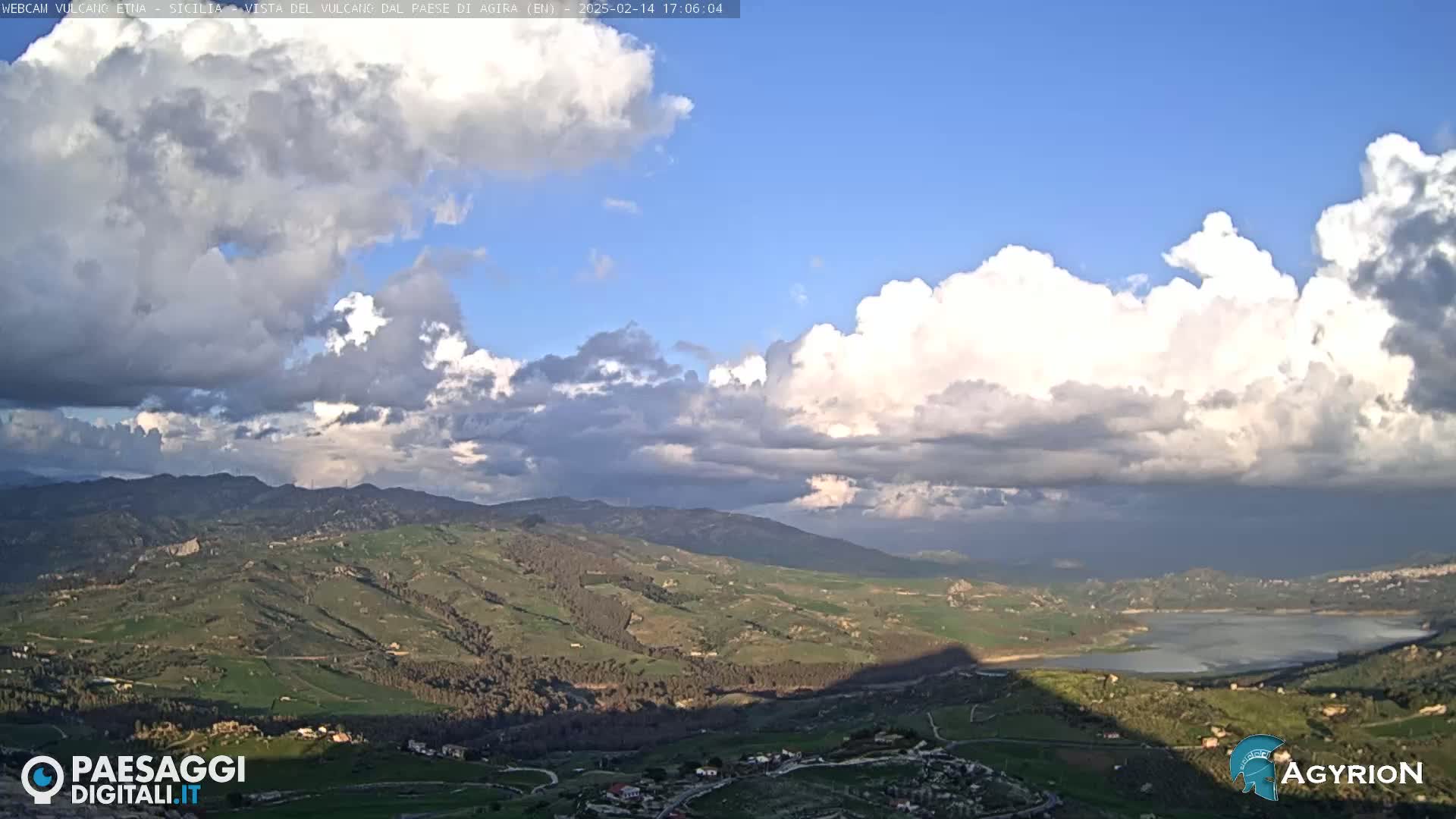 A partly cloudy sky overlooks a vast, hilly landscape containing a lake, with shadows cast across the valley.