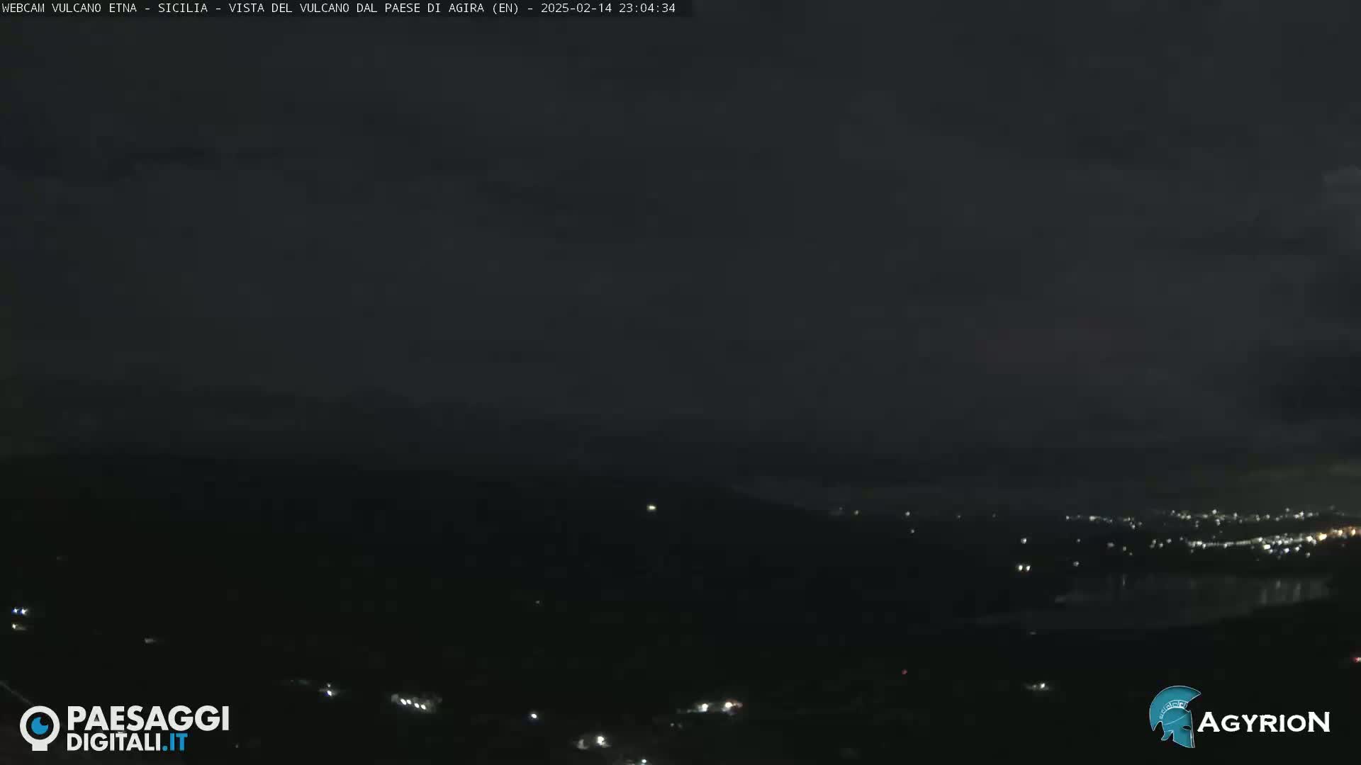 A nighttime long shot of a dark, mountainous landscape with a few scattered lights visible in the distance under a mostly clear, dark sky.
