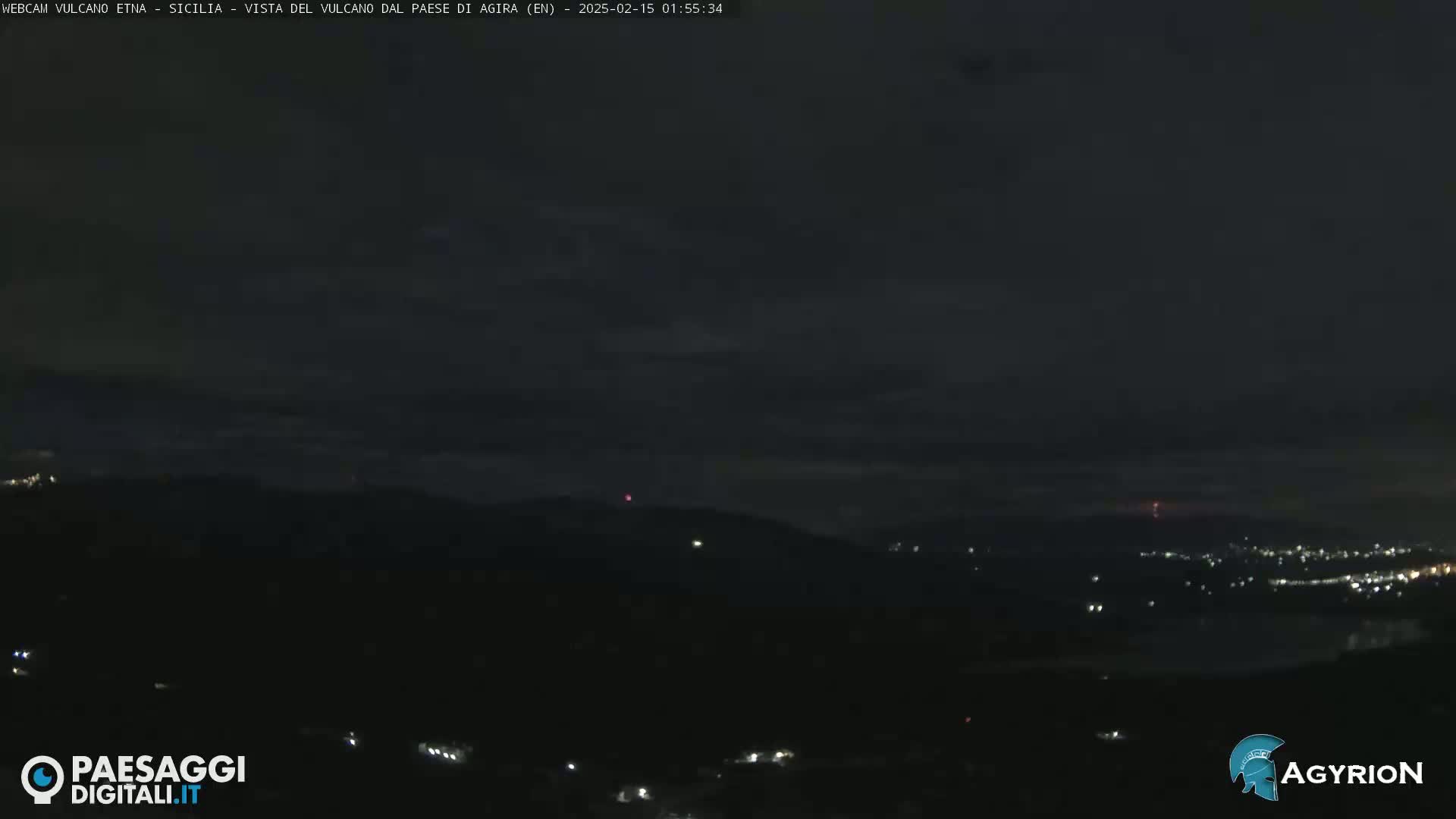 A nighttime long shot of a dark, hilly landscape with scattered lights and a faint reddish glow on the horizon under a mostly clear sky.